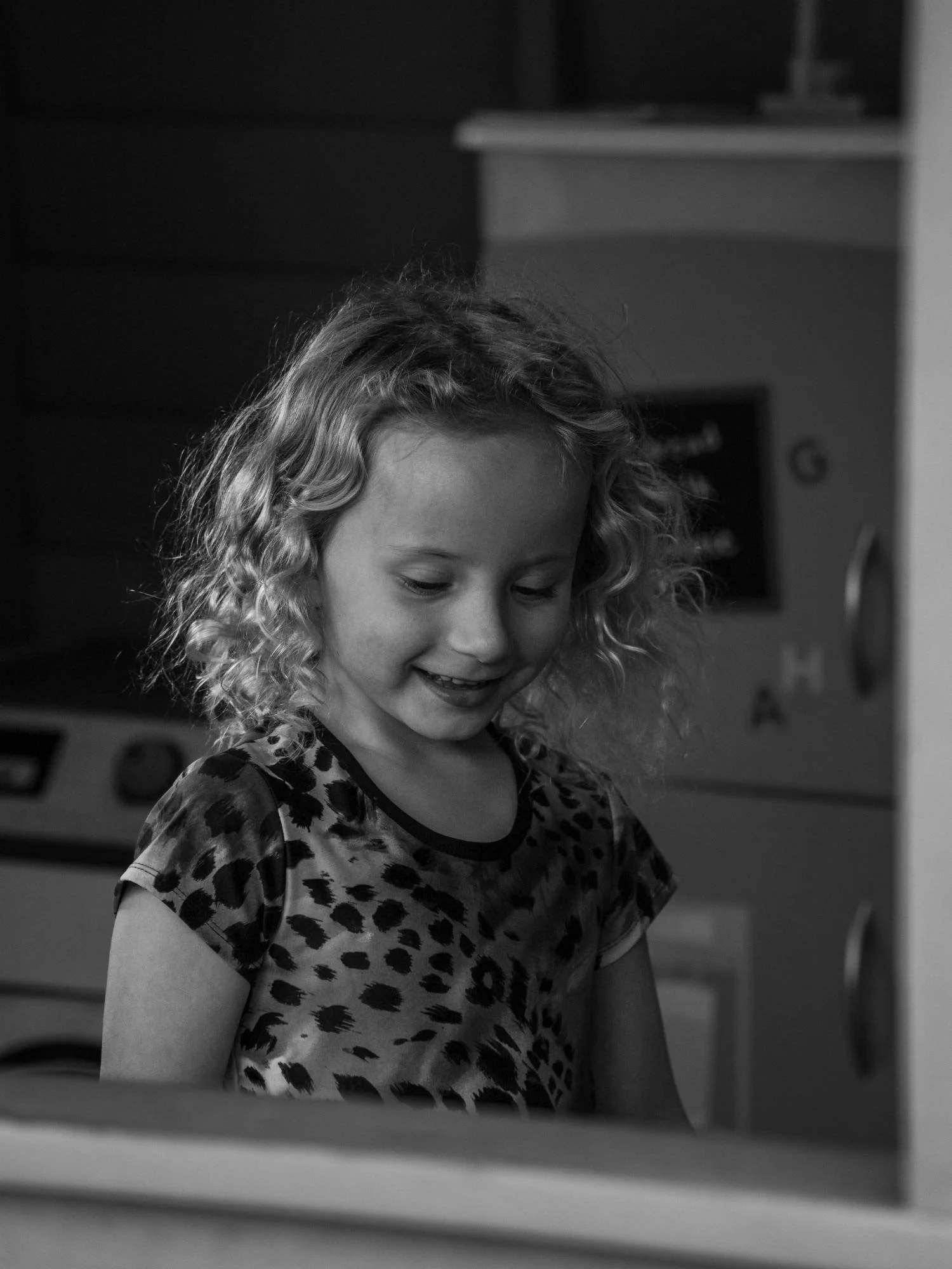 Photograph of a girl with blonde, curly hair, smiling through the window of a cubby house