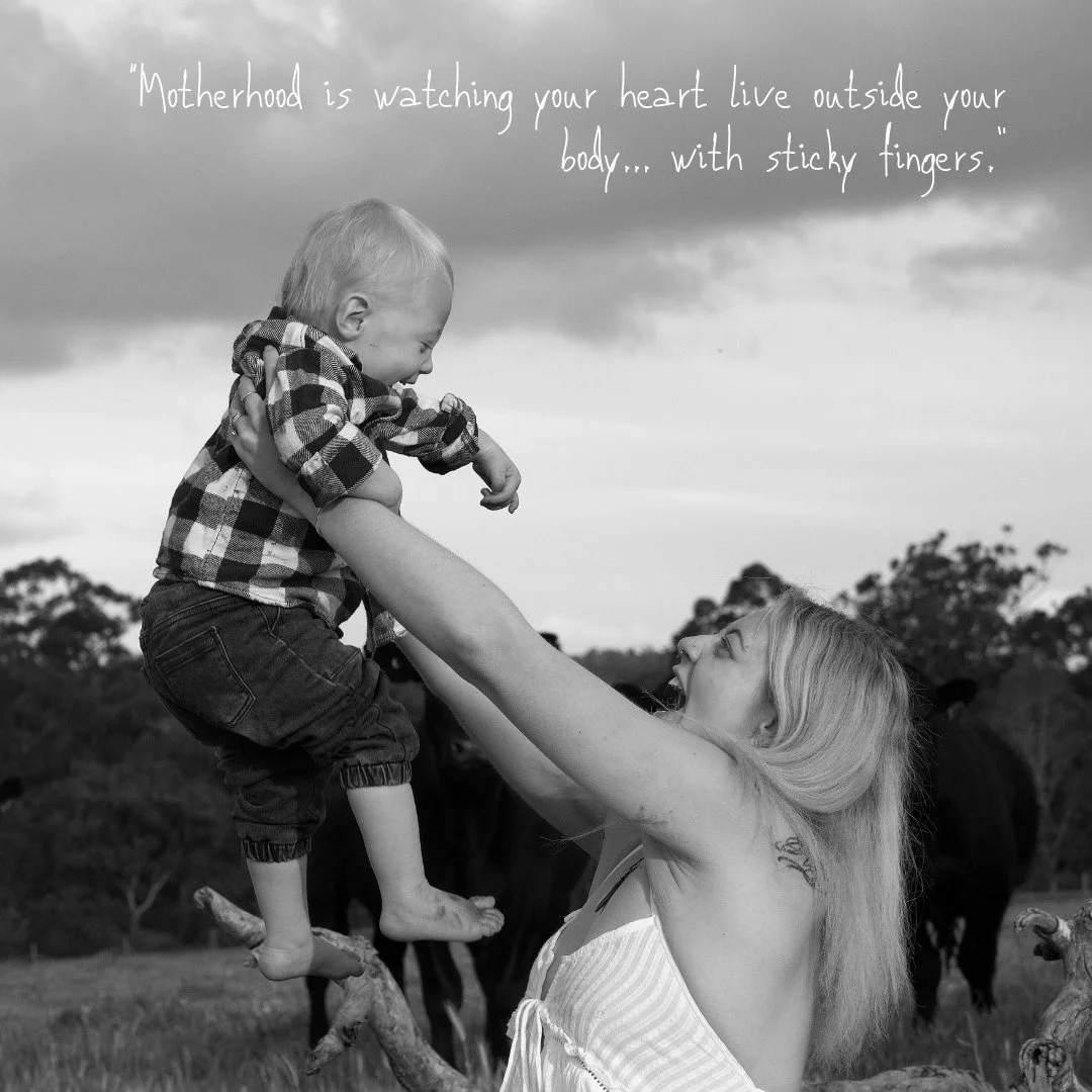 Black and white image of a mother lifting her son in the air. caption reads: Motherhood is watching your heart live outside your body... with sticky fingers.