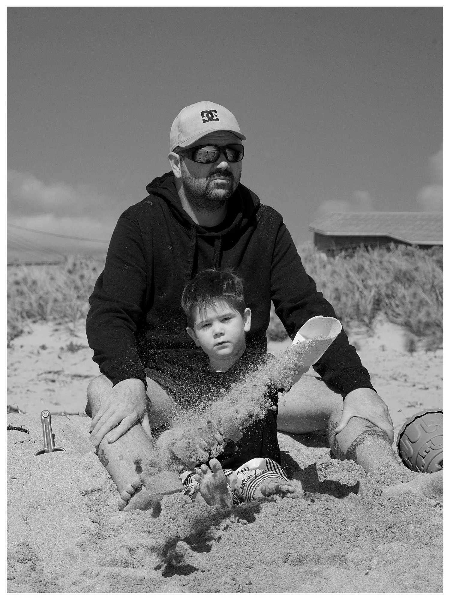 A man and a young boy playing with a shovel on the sand at the beach. The man is sitting with his legs spread, wearing a black hoodie, sunglasses, and a baseball cap. The boy is sitting in front of him, with the shovel raised, and sand flying in the 
