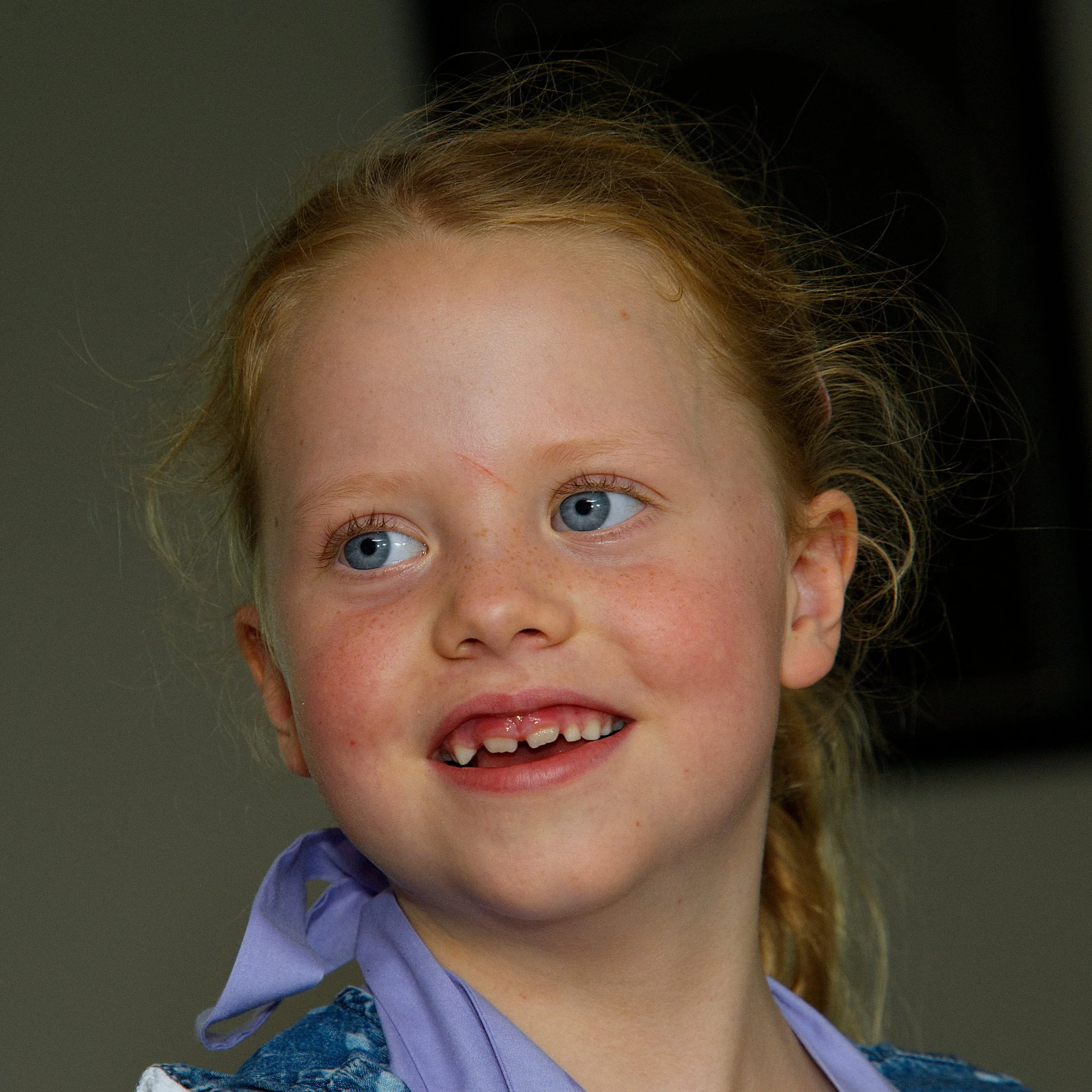 Close-up of a young girl with red hair, blue eyes, freckles, and a missing tooth, smiling and wearing a bluish top and purple necktie.