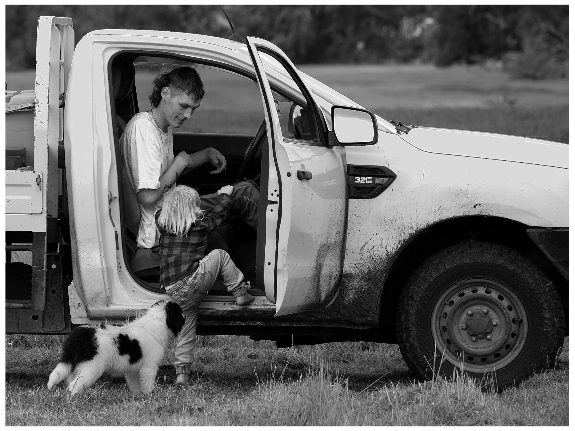 A man and a young child sitting in the open door of a pickup truck outdoors. A dog is near the child's feet. The man and child are engaging with each other, and the scene is black and white.
