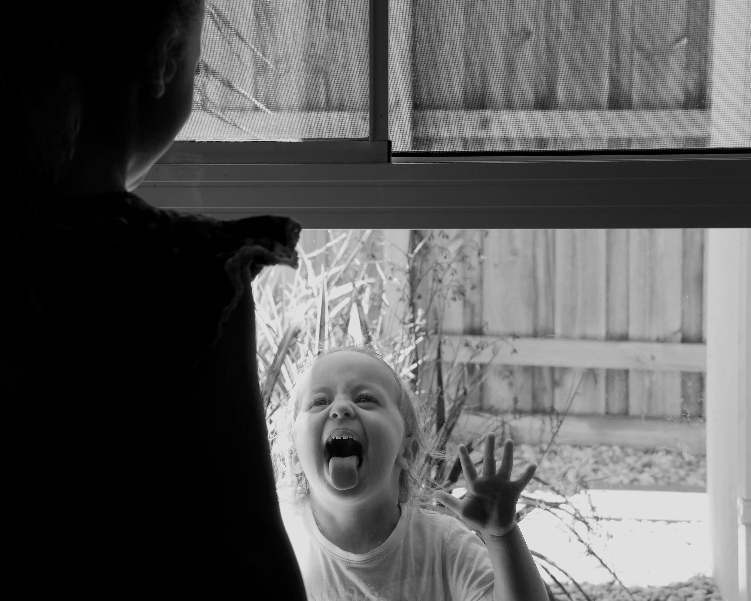 A young girl is standing outside a glass door, sticking her tongue out and raising her hand, while a person inside the house looks on. The scene is in black and white.