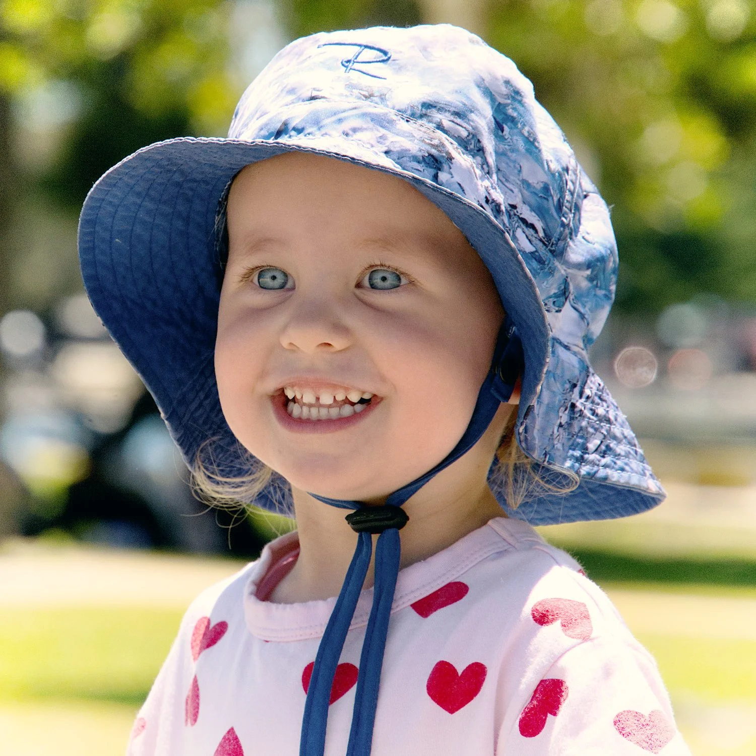 A smiling young girl with blue eyes wearing a blue and white patterned sun hat and a white shirt with red hearts, standing outdoors with trees and green grass in the background.