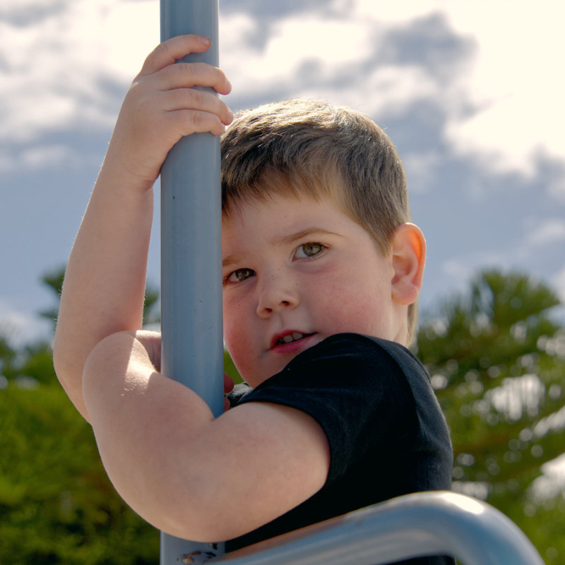 A young boy with brown hair looks shyly from behind a blue pole at a playground