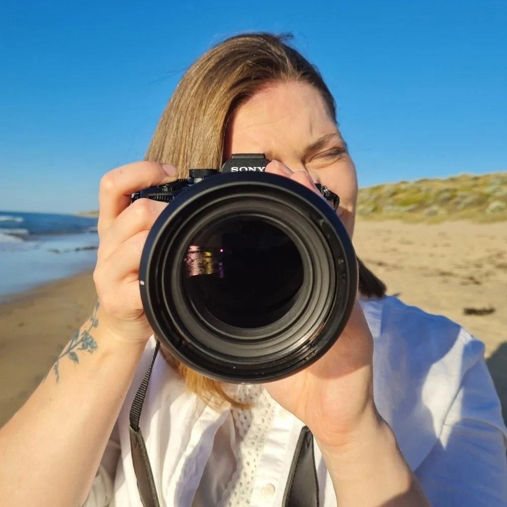 Stacey Potter taking a photo at Halls Head beach with camera, sandy shore and blue sky in background.