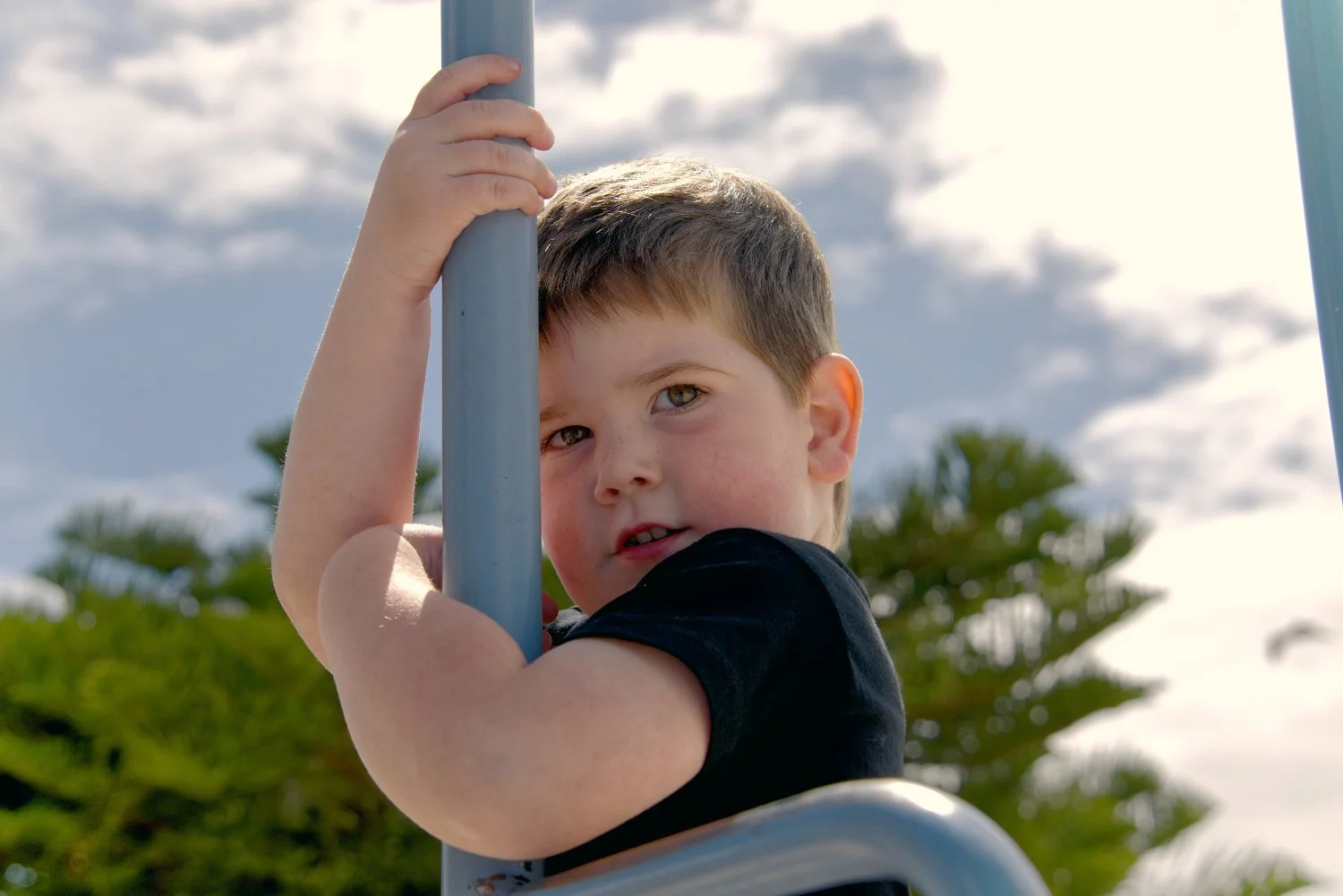 Young boy with short brown hair and fair skin looking over a metal railing outdoors against a backdrop of blue sky, clouds, and green trees.