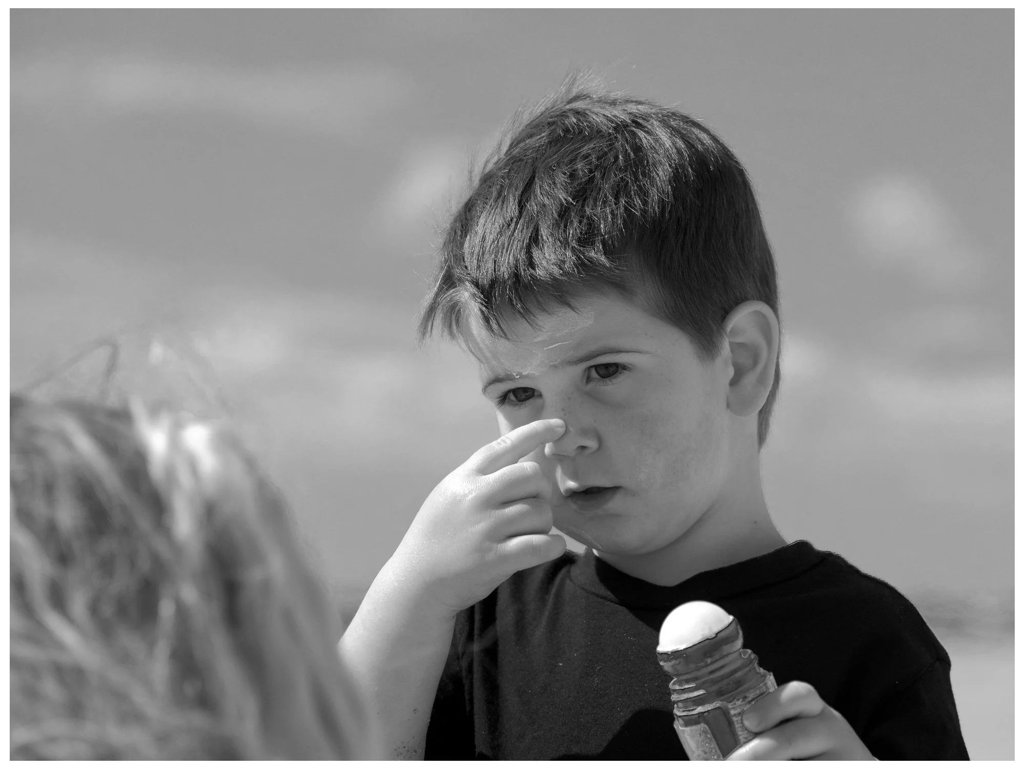 A young boy with short dark hair holding a sunscreen bottle, touching his nose.