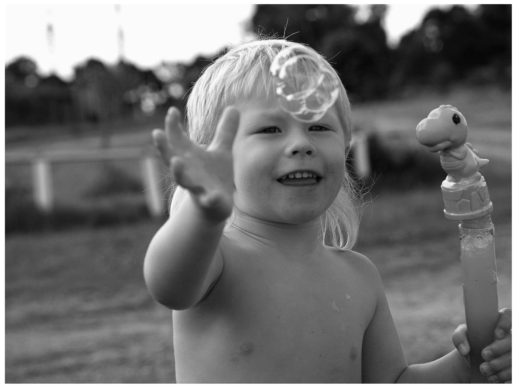 A young child with light hair playing outdoors, reaching towards a soap bubble in a backyard.