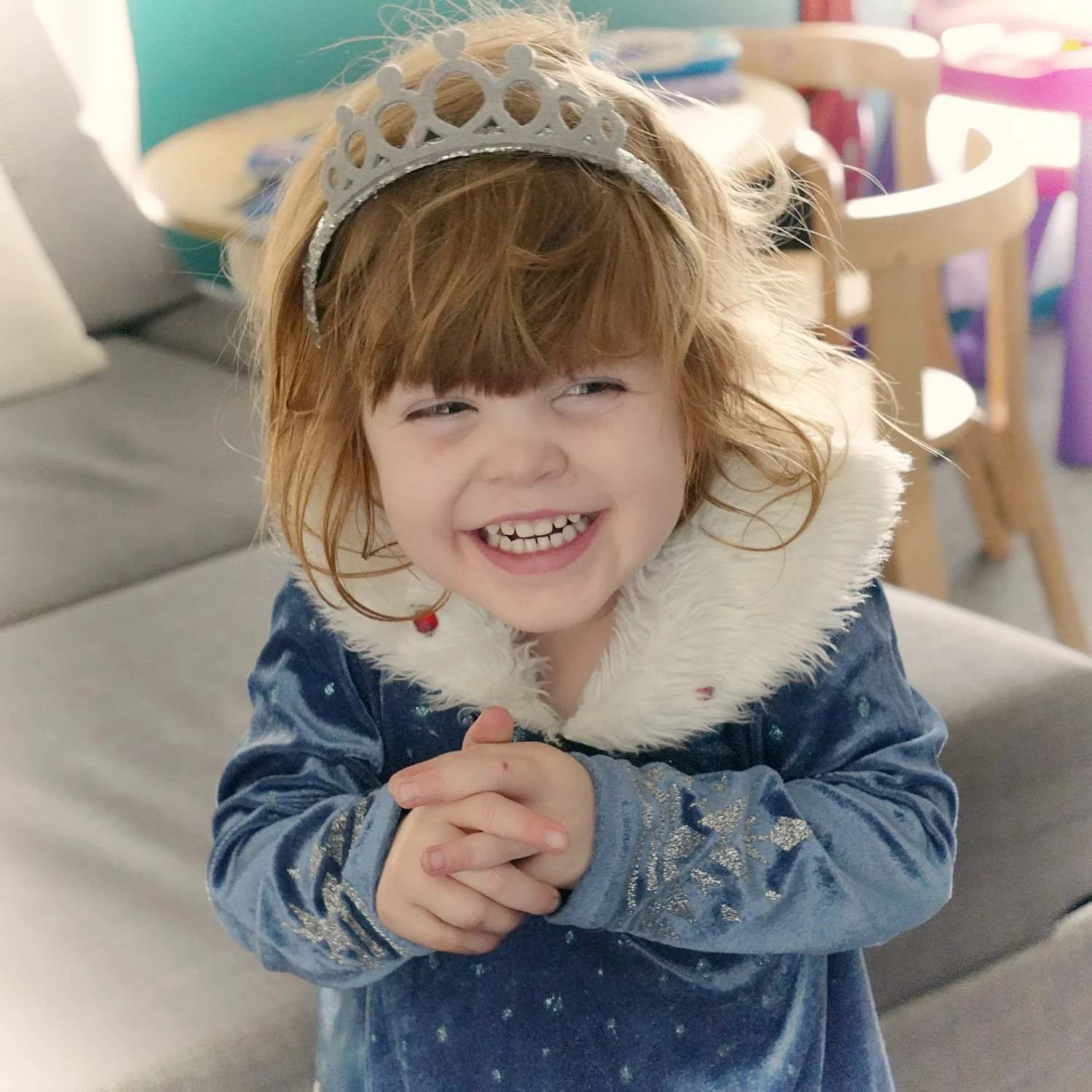 A girl in a tiara and blue princess dress clasps her hands and smiles at the camera