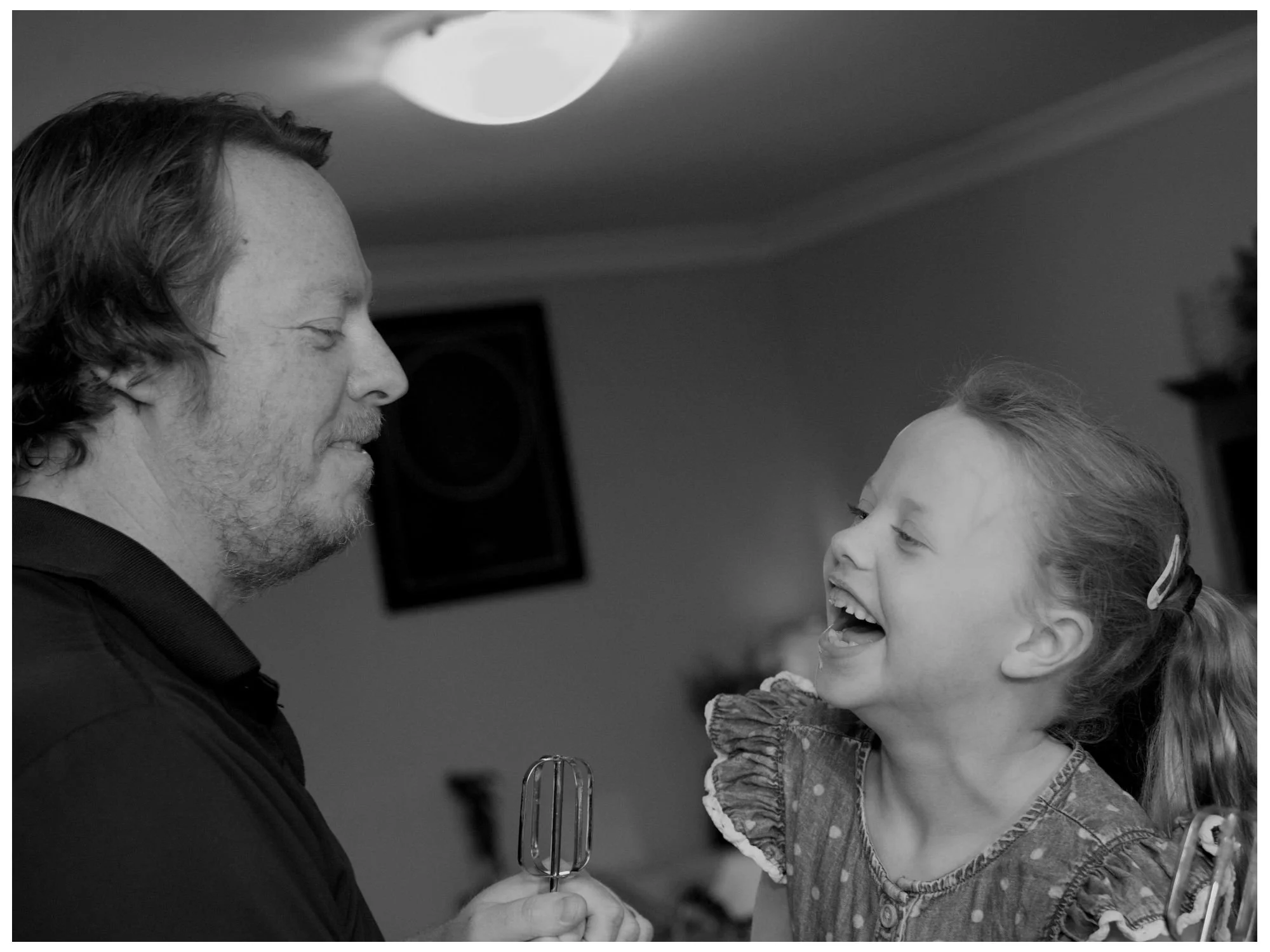 A man and young girl sharing a joyful moment in a kitchen, with the girl laughing and the man holding a hand mixer.