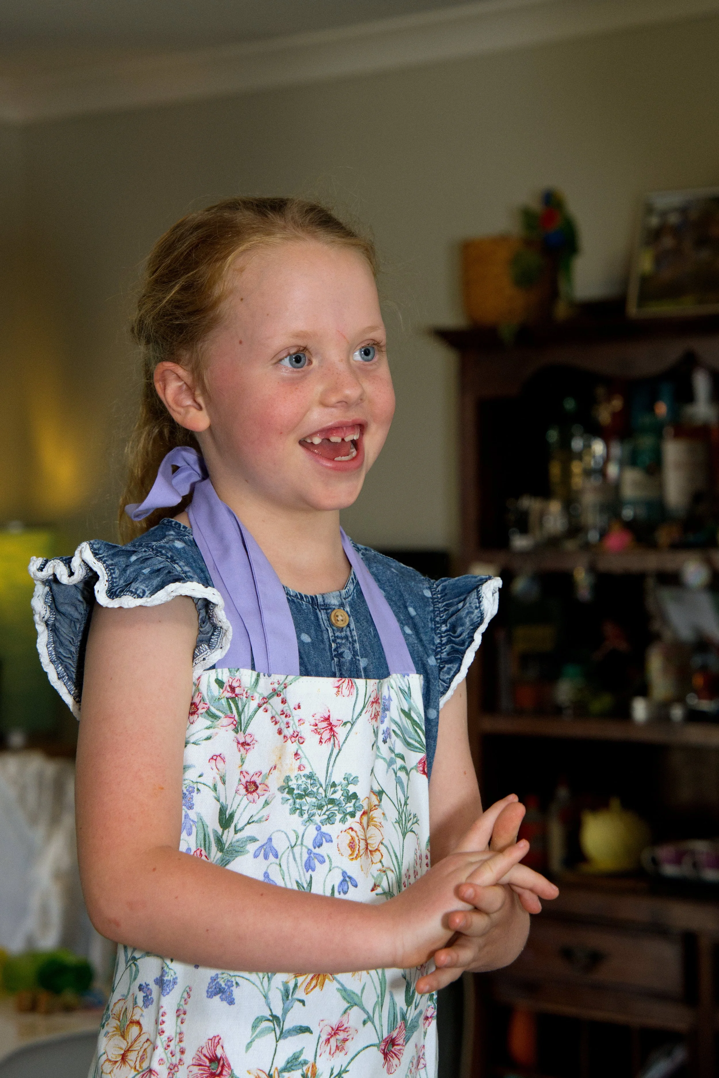 A young girl with red hair, blue eyes, smiling, wearing a blue dress with ruffled sleeves and a floral apron, standing in a kitchen.