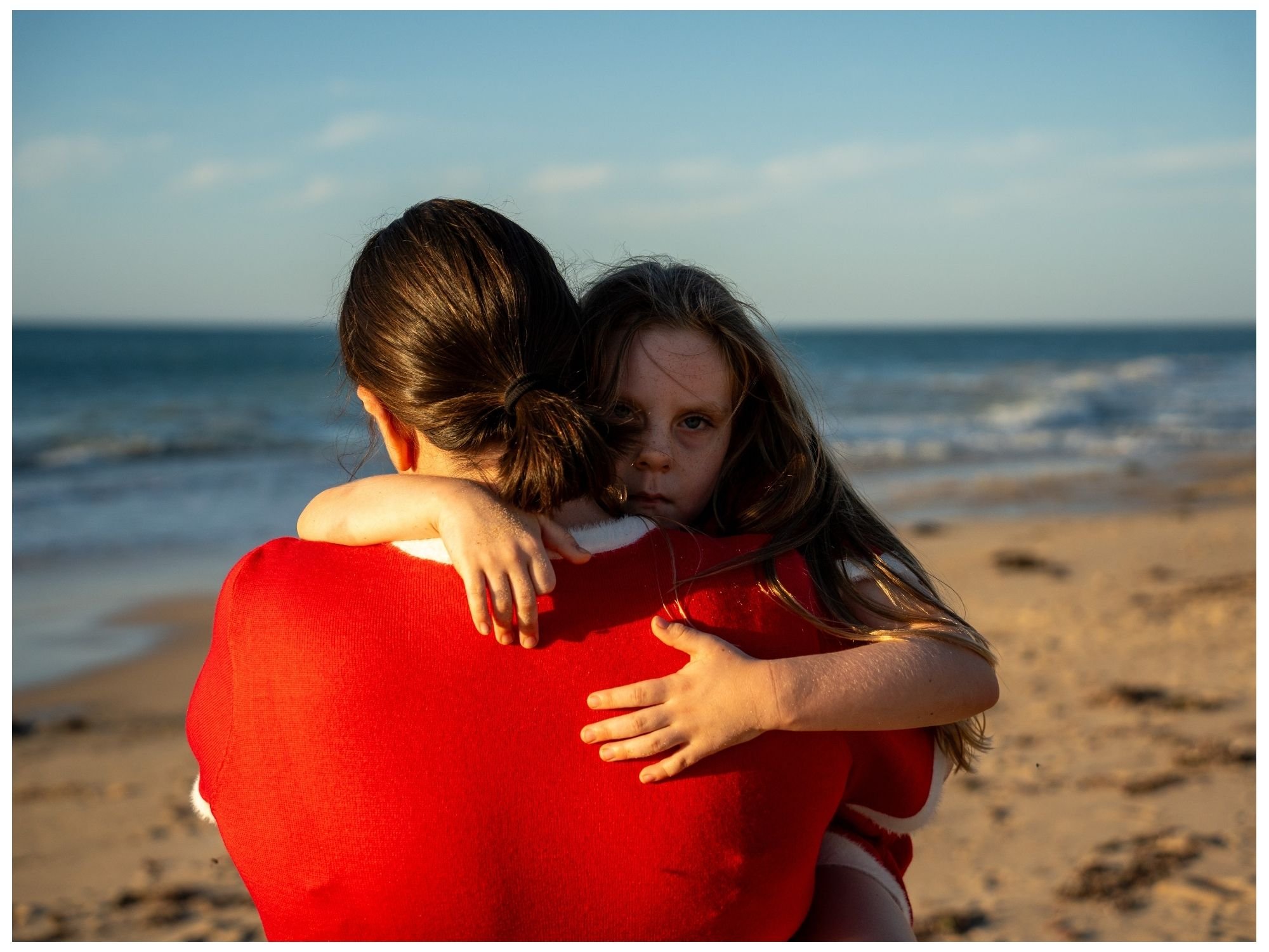 A woman is hugging a young girl at the beach, with long, disheveled hair, and is staring directly at the camera. The woman’s back is to the camera, and she is wearing a red garment. The ocean and sky are visible in the background.