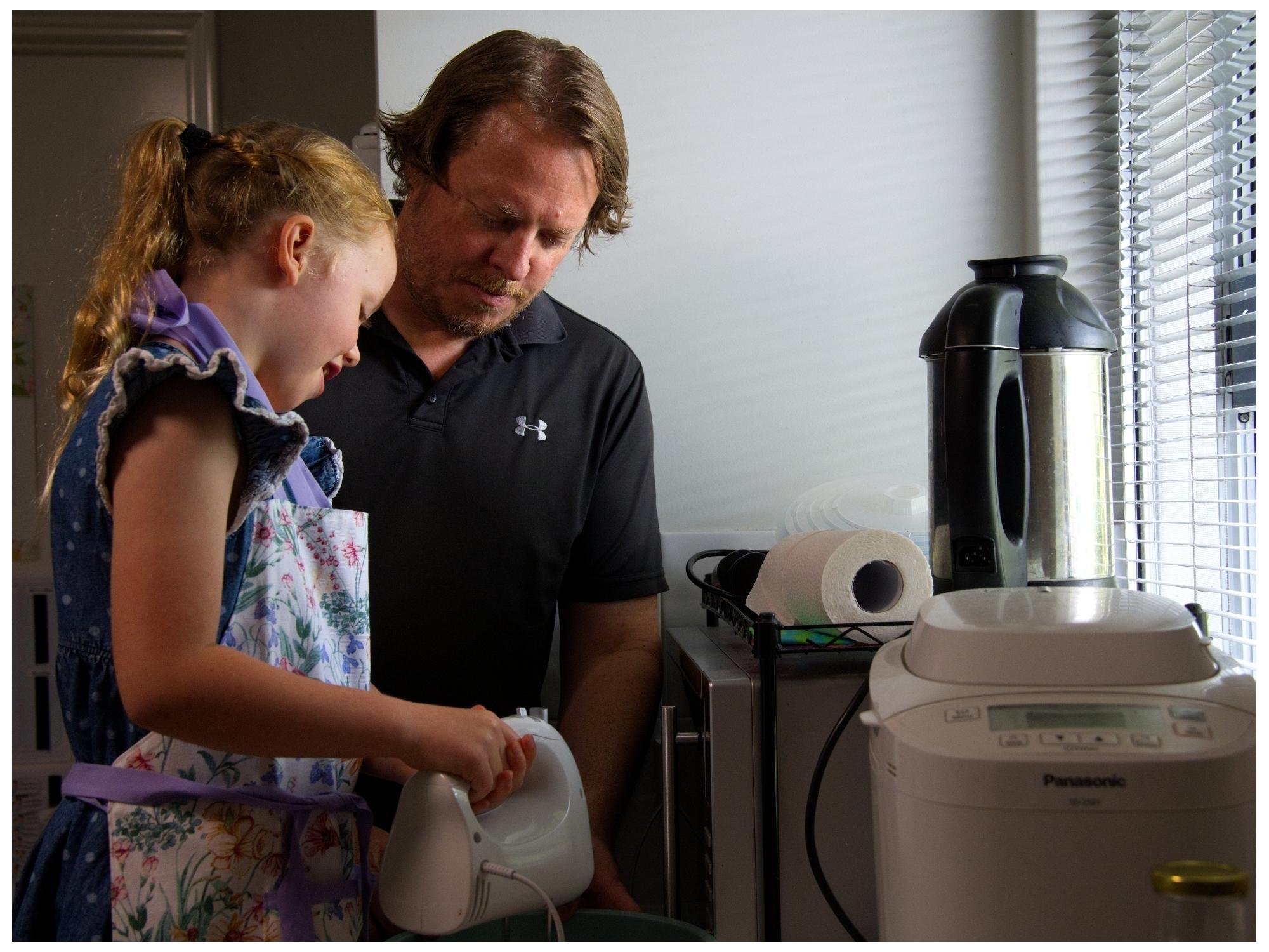 A man and a young girl are in a kitchen, looking at an electric mixer the girl is holding. The girl is wearing a floral apron and purple dress, and the man is wearing a black polo shirt. Kitchen appliances and paper towels are visible on the counters