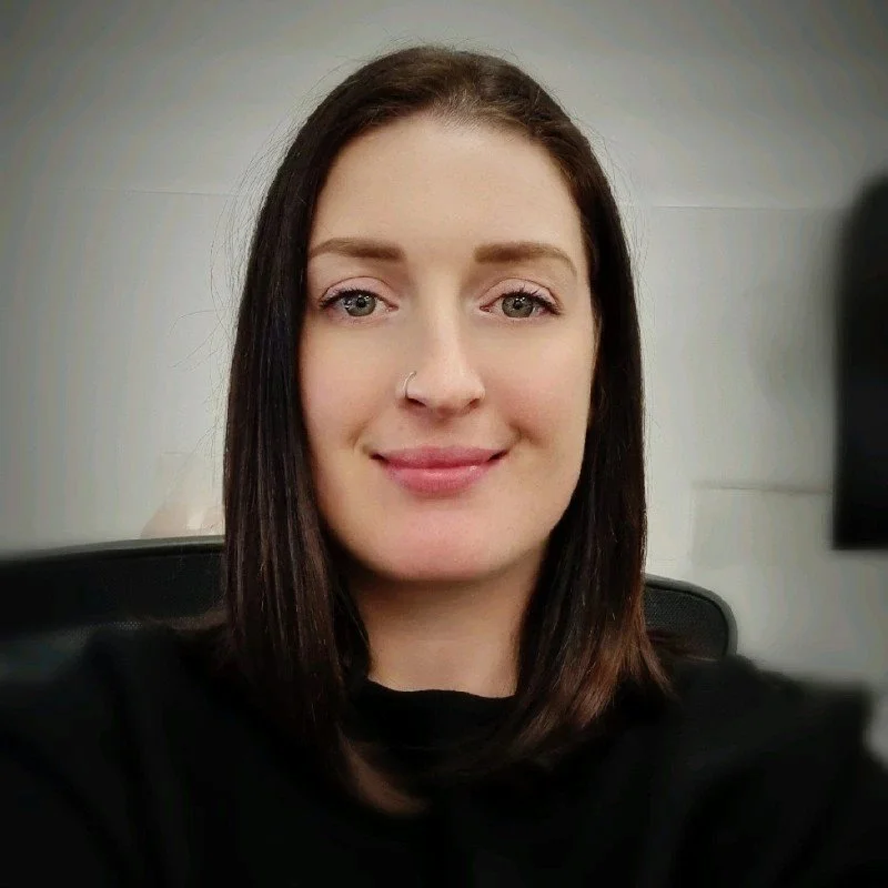 A woman with straight brown hair, wearing a black top, smiling at the camera in a well-lit room.