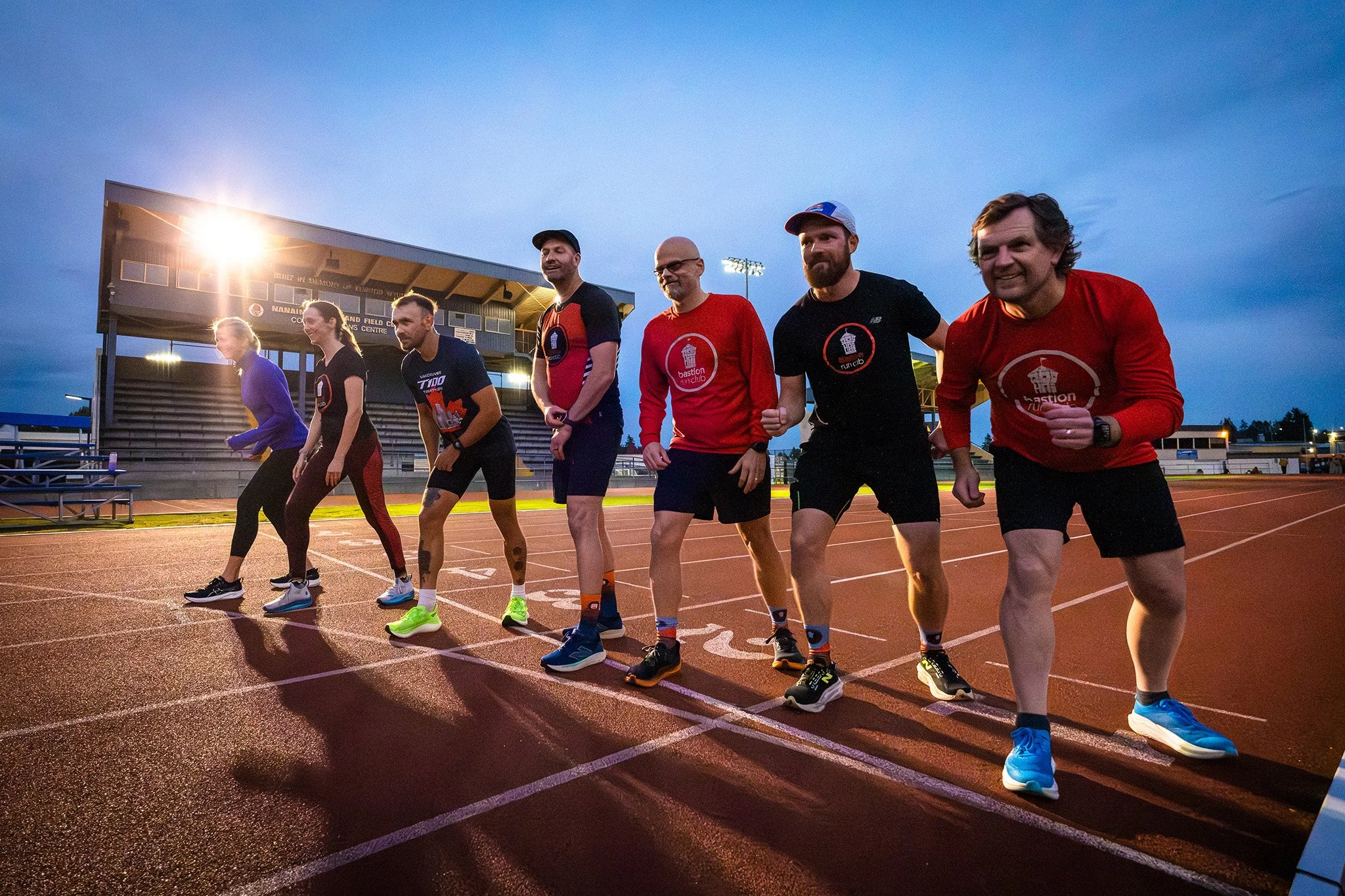 A group of runners on a track at dusk, in sportswear, preparing for a race at a stadium with bright lights.
