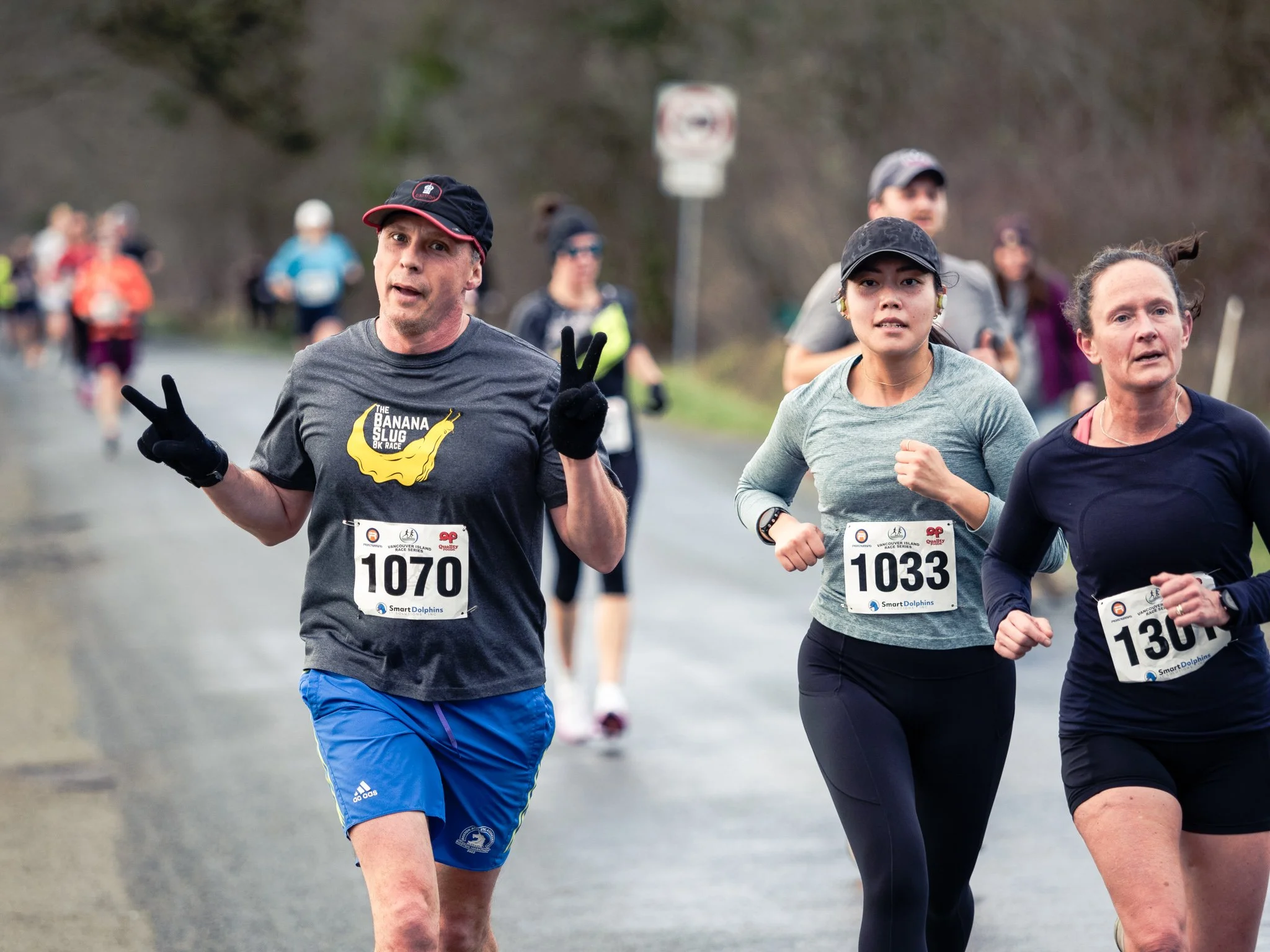 A group of people participating in a running race outdoors, wearing athletic clothing and race bibs, with trees and a blurred background.