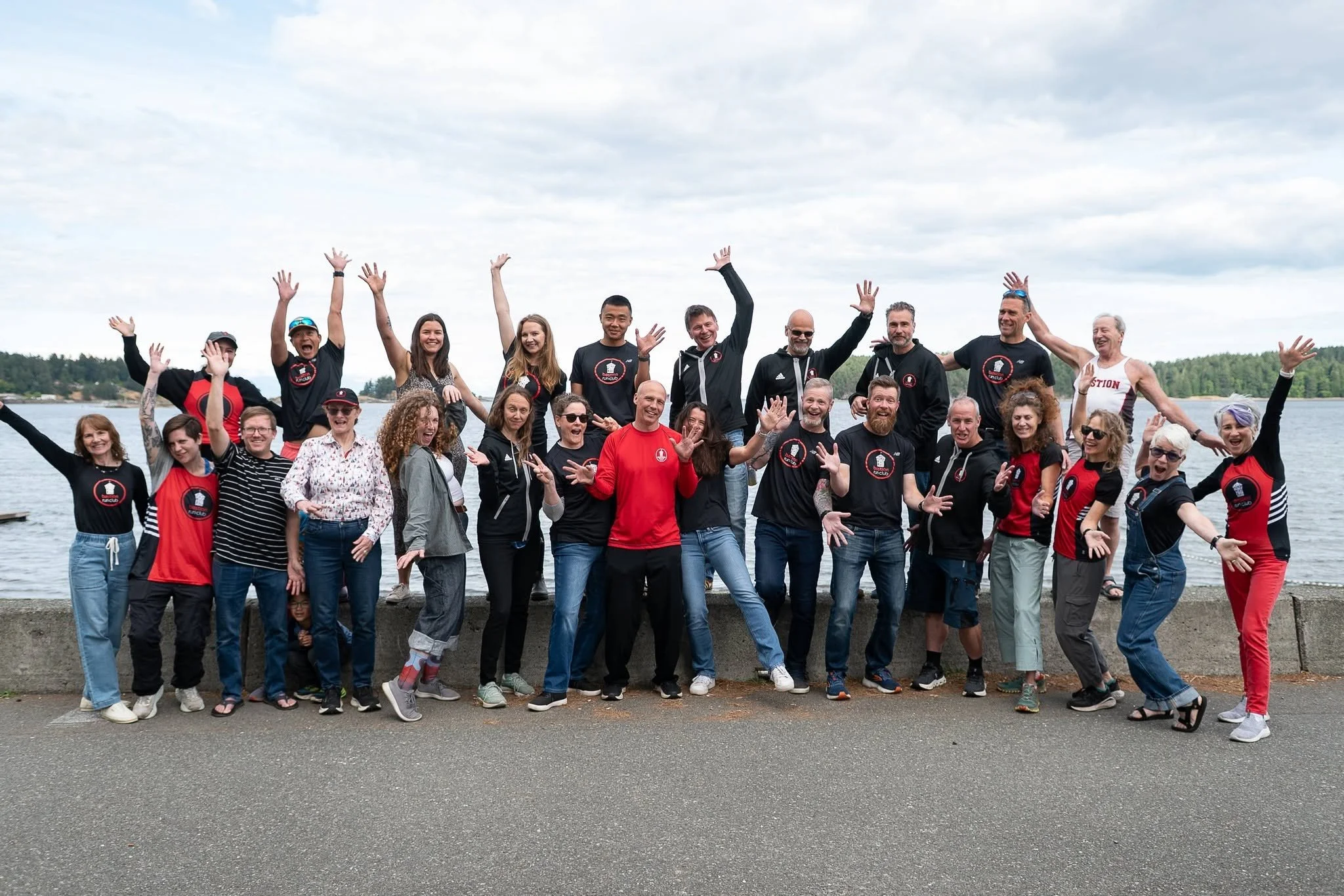 A large group of diverse people smiling and posing by the water during daytime, with some wearing Bastion Run Club branded clothing
