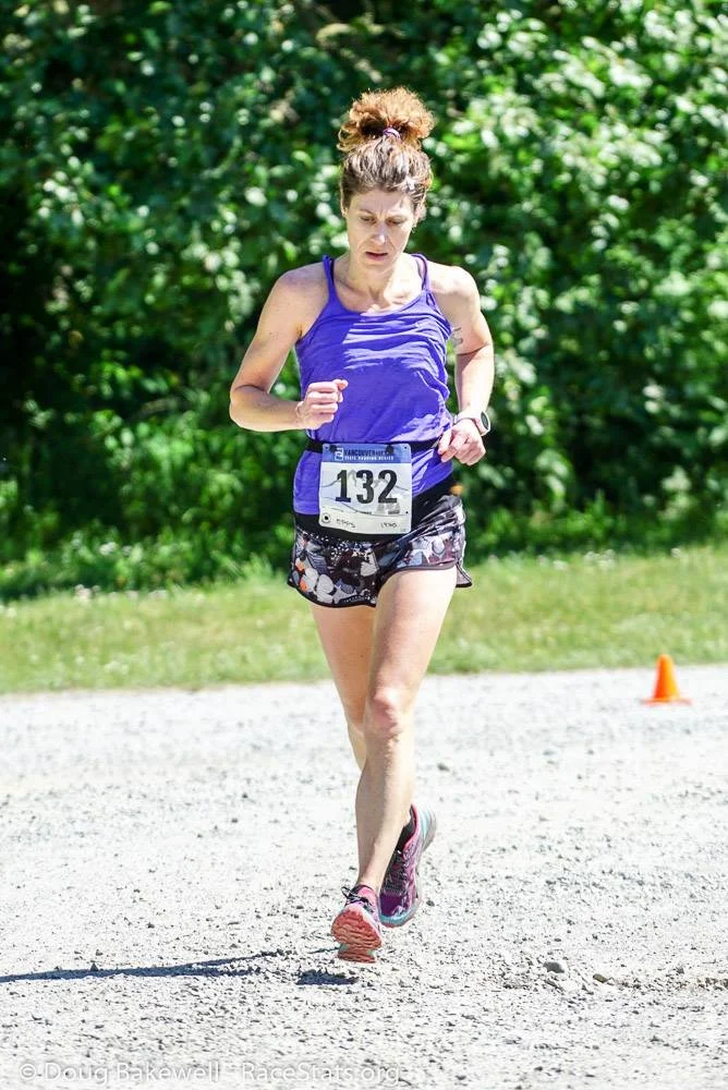 A woman running outdoors wearing a purple tank top, patterned shorts, and pink running shoes, with a race number 132 attached to her waist, and an orange cone in the background on a gravel path surrounded by green trees.