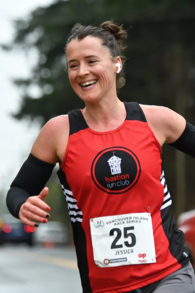 A woman running in a race, wearing a red and black sports top with 'bastion run club' logo, race bib number 25, and wireless earbuds, smiling as she runs outdoors.