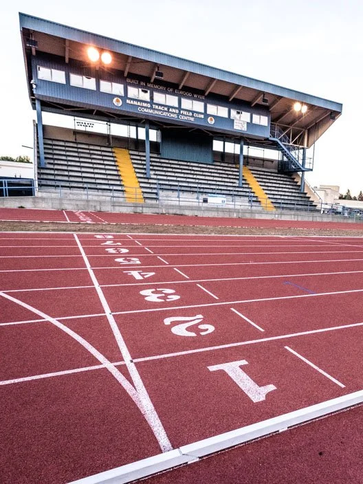 A view of a running track at a stadium with marked lanes numbered 1 through 6 in white. The background shows bleachers and a building with signage for Nanaimo Track and Field Club, with curtains and lighting. The sky is clear, indicating outdoor stadium conditions.