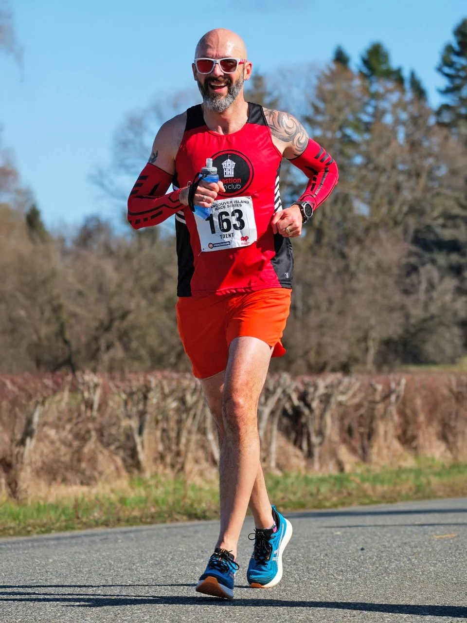 A man running outdoors during a race, wearing a red sleeveless shirt, orange shorts, and blue running shoes, with a race bib numbered 163 and sunglasses, smiling with trees and clear sky in the background.
