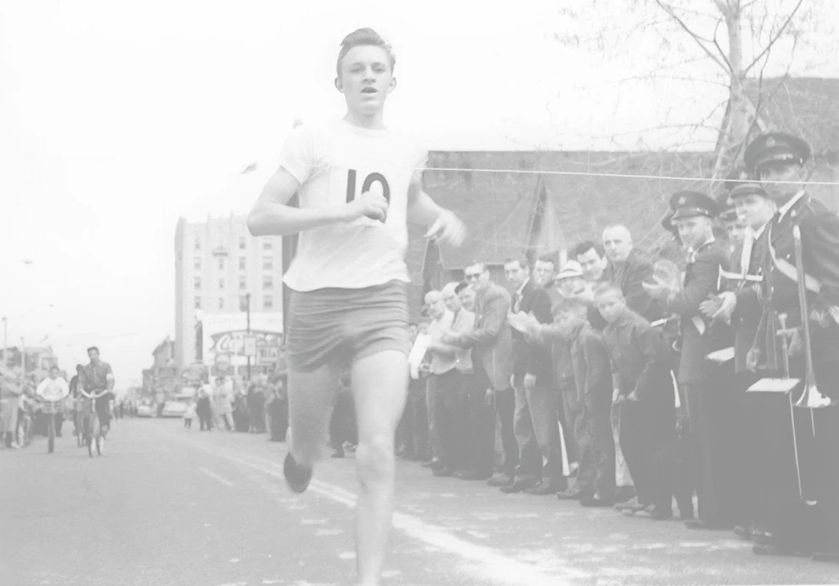 A young male runner participating in a race on a city street, with spectators and officials watching along the side of the road, some clapping.
