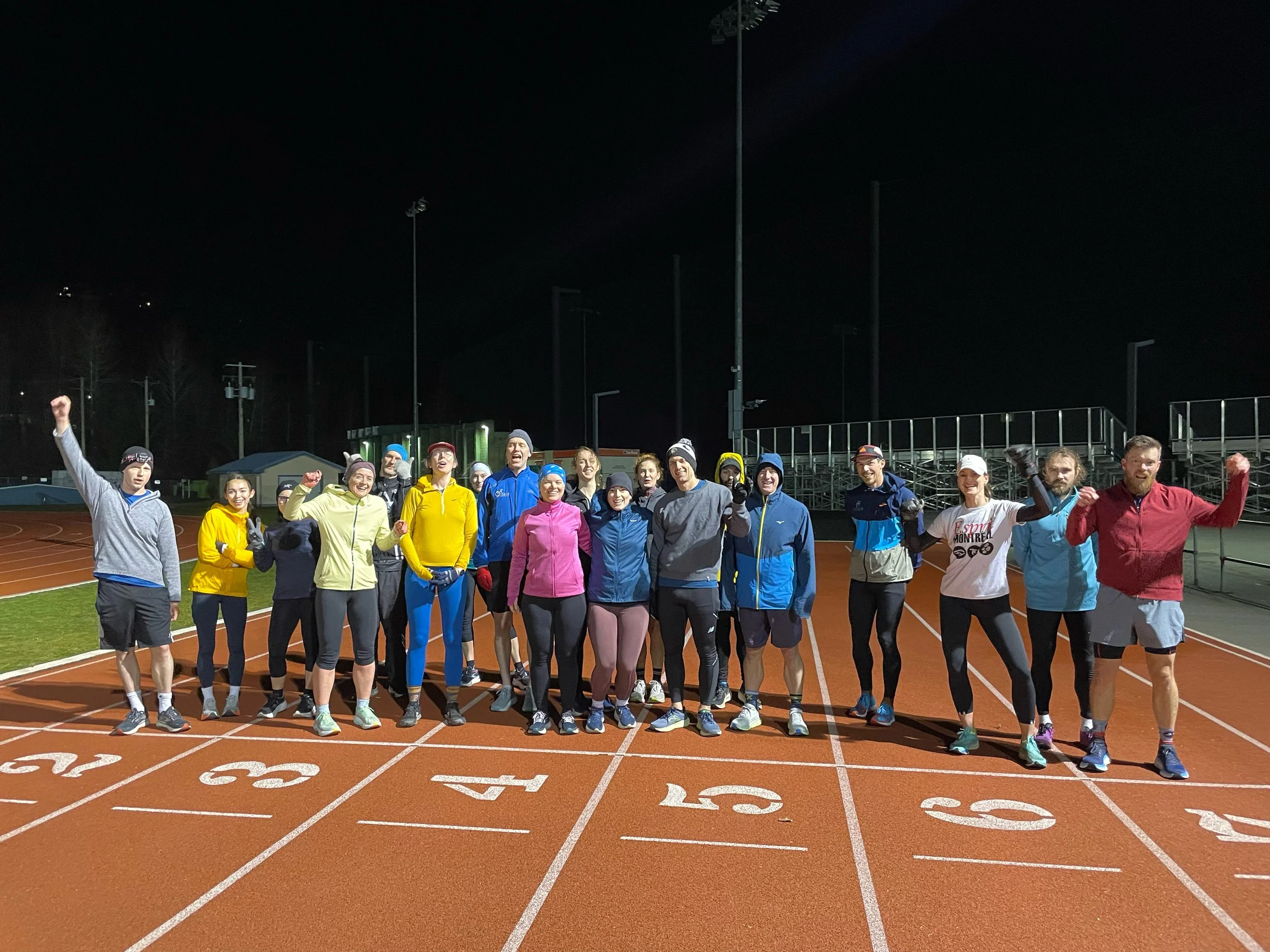 A group of runners in athletic clothing and winter hats, raising their fists and smiling on a red running track at night, with tall stadium lights and bleachers in the background.