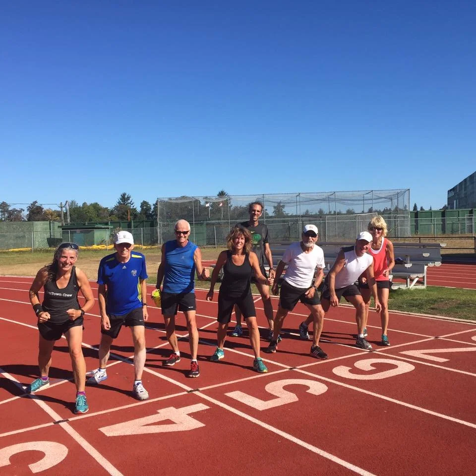 A group of nine people, men and women, in athletic clothing, are gathered at the starting line of a running track, ready for a race, under a clear blue sky.