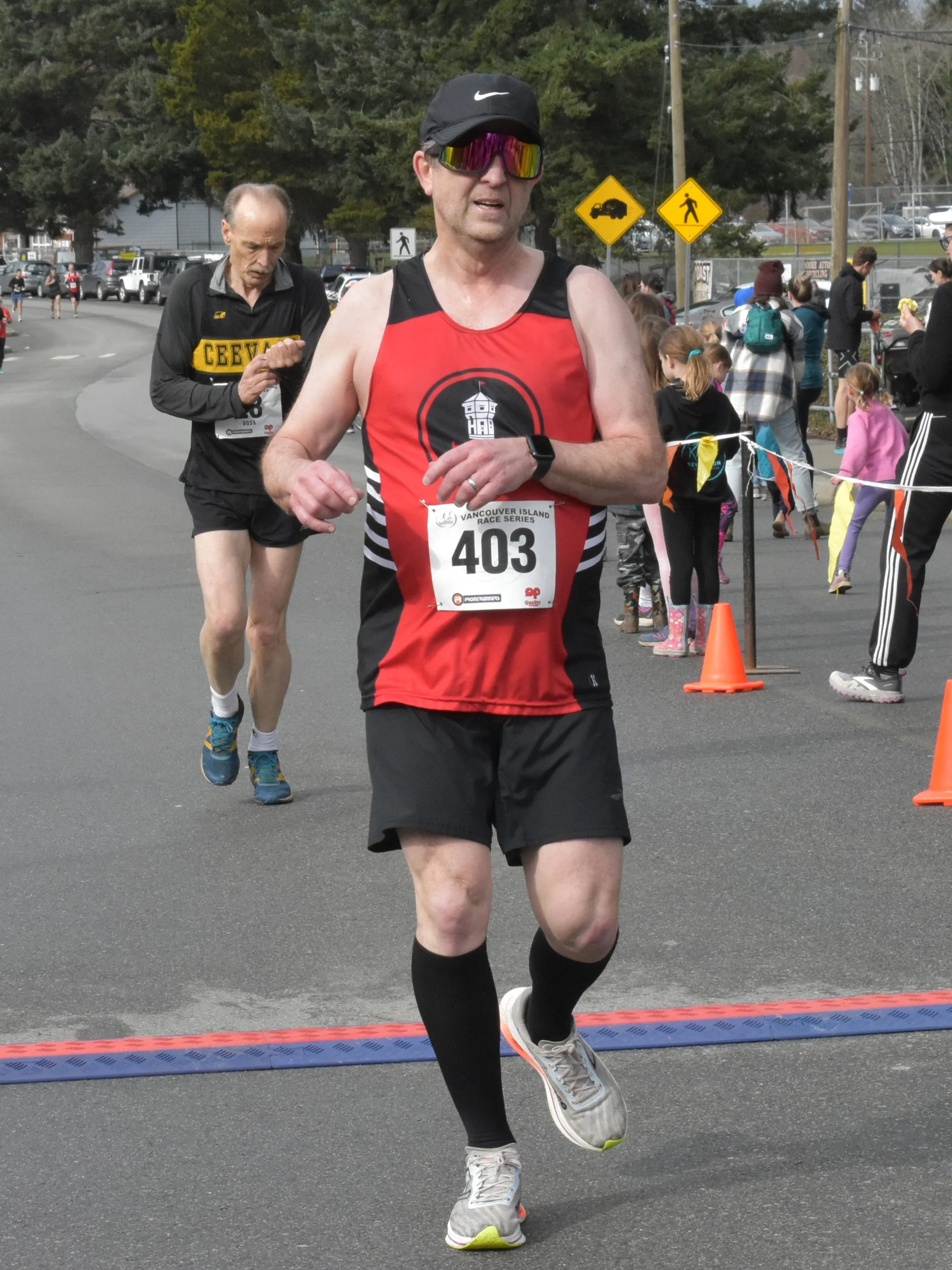 A man with a race bib number 403 crossing the finish line in a race, wearing a black cap, reflective sunglasses, and a red and black running outfit. There are spectators and other runners in the background.