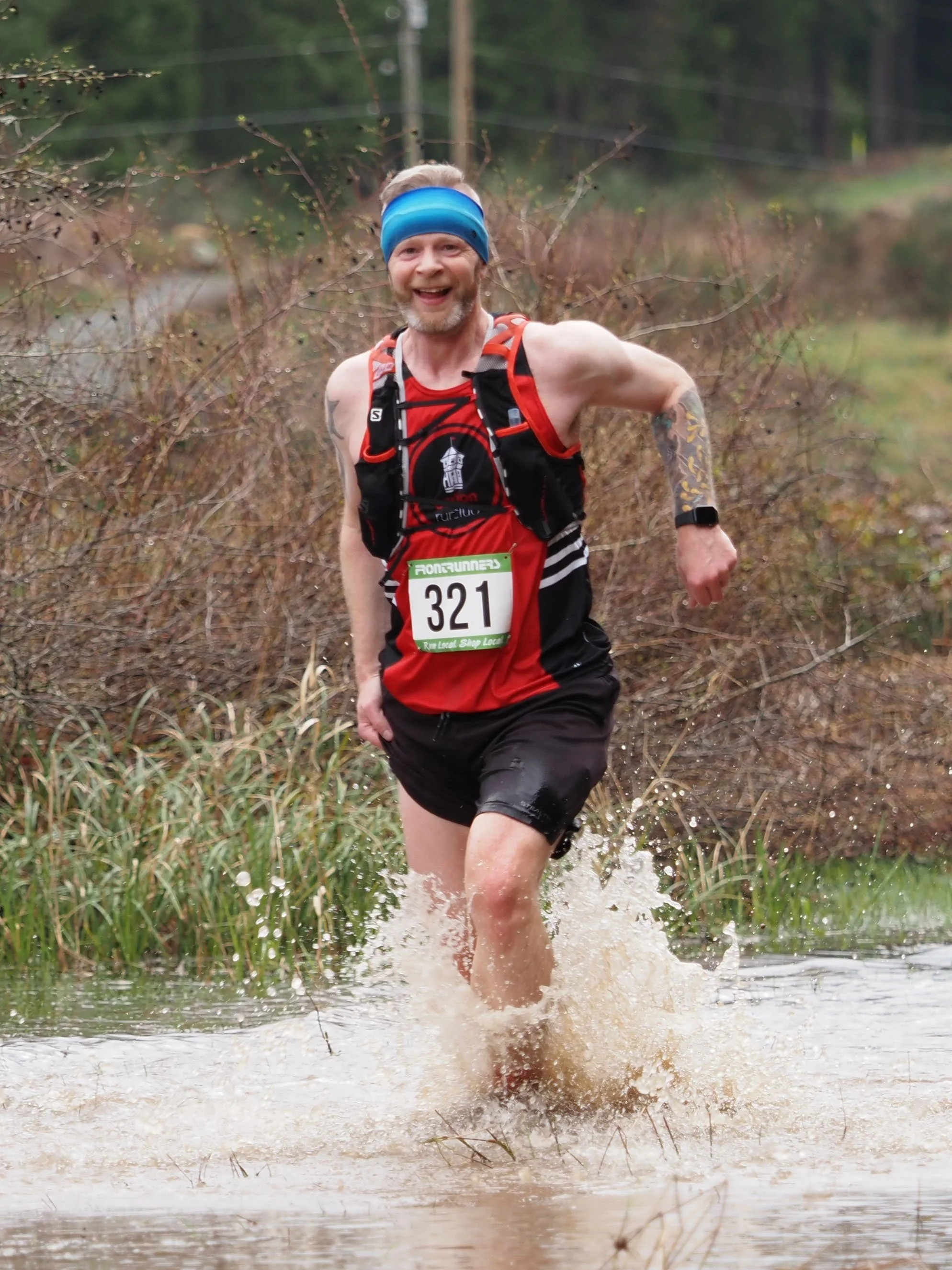 A man running through a shallow water crossing during a trail race, wearing a red and black running outfit, bib number 321, a blue headband, a hydration pack, and a smartwatch.