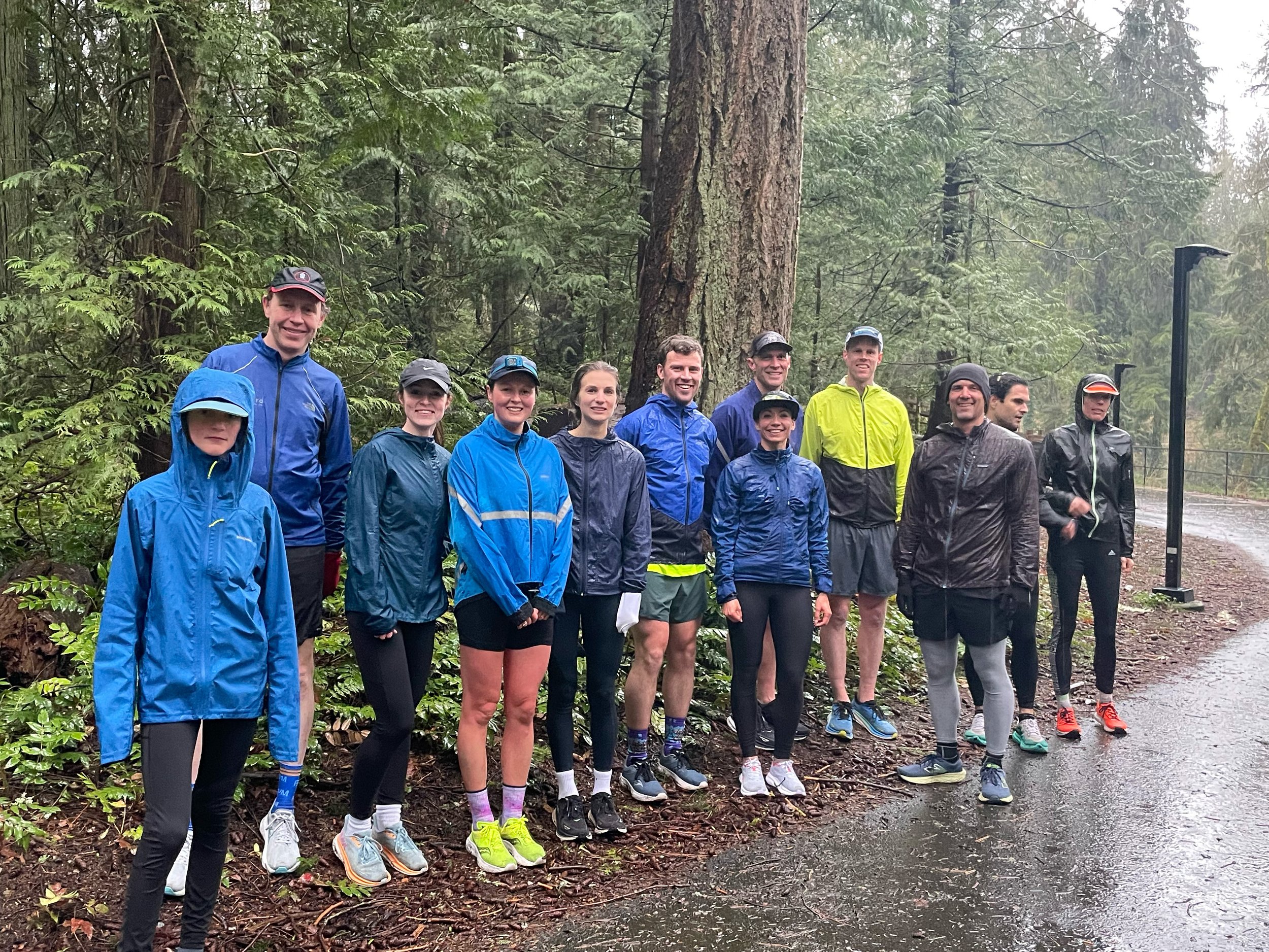 A group of about 12 runners standing together in a forest, wearing athletic clothing and rain gear, with tall trees and a path in the background on a rainy day.