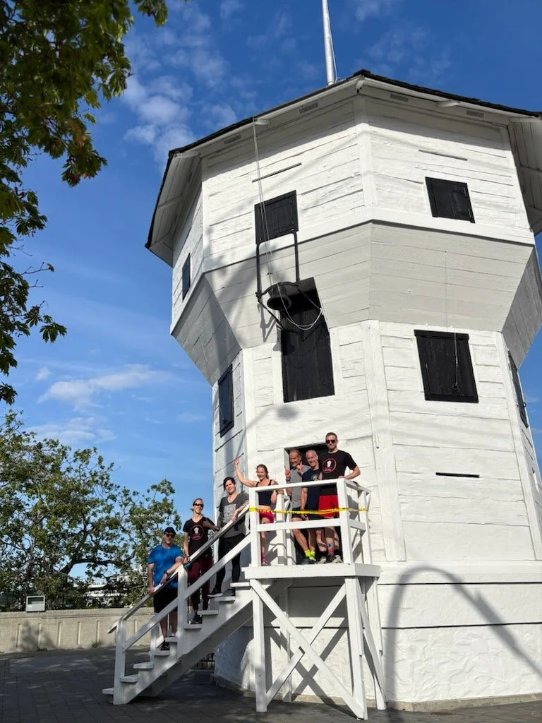 Group of six people standing on a small wooden deck in front of Nanaimo's historic Bastion building, clear blue sky in background.