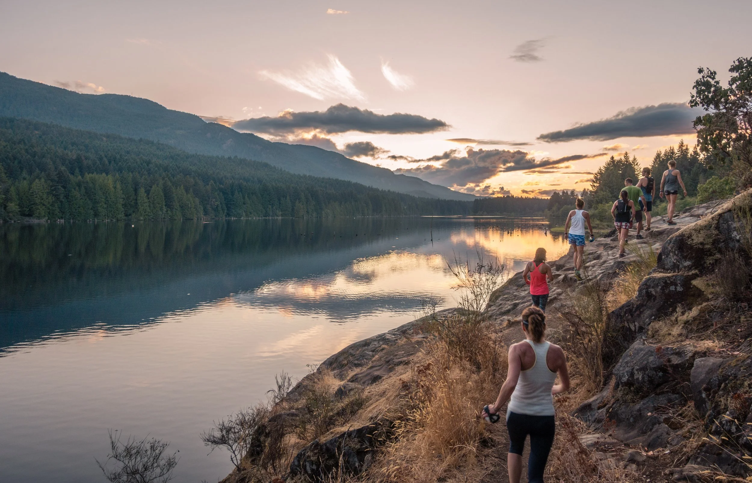 A group of people walking on a rocky trail beside a calm lake during sunset with mountains and clouds in the background.