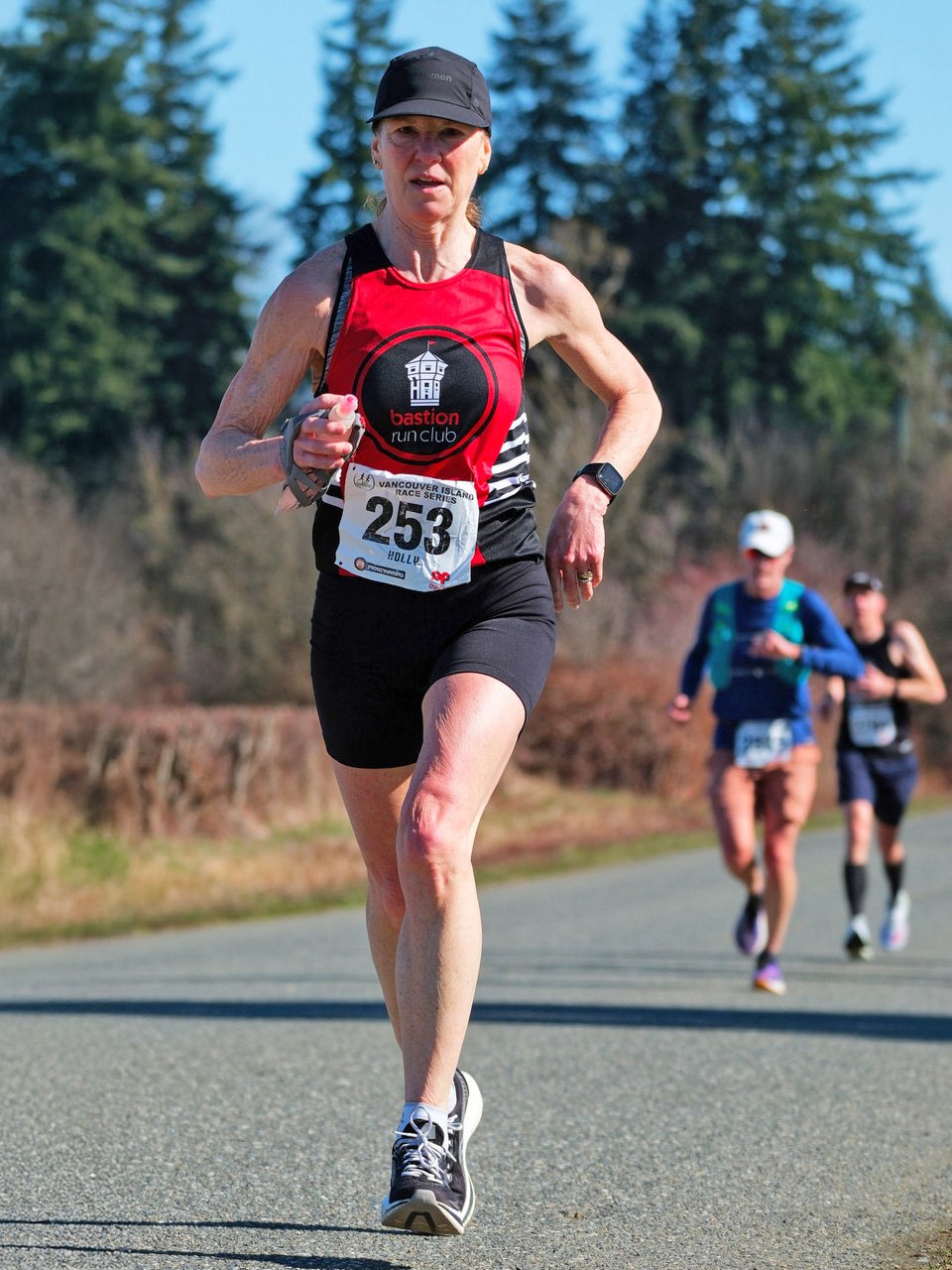 A woman running in a race, wearing a black and red athletic outfit with a bib number 253, on a rural road with a background of trees and other runners.