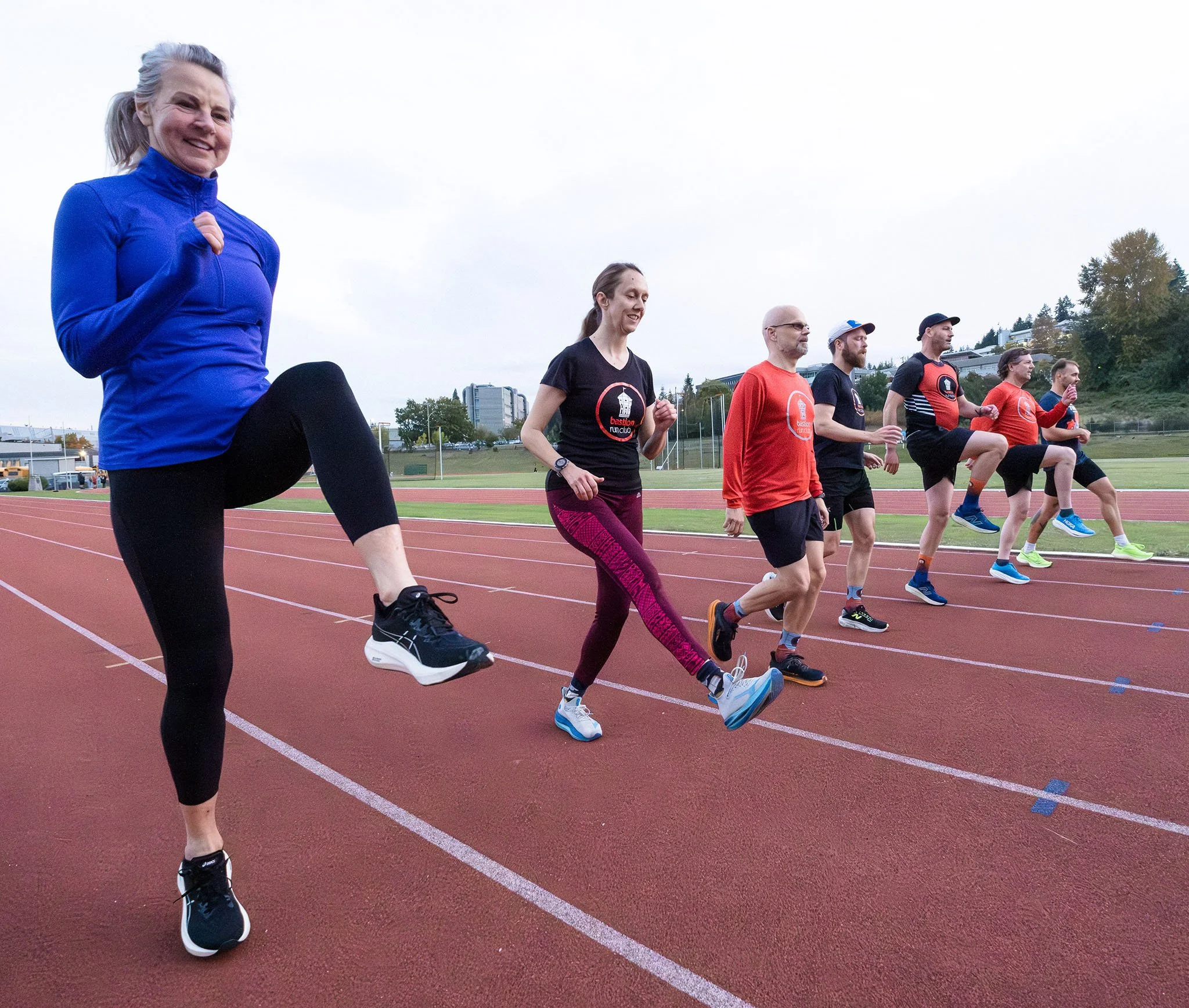 A group of people jogging on a running track during daytime, with a taller woman in a blue jacket leading in the foreground.