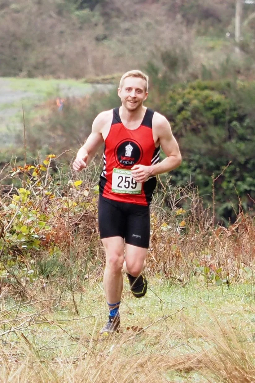 A man running outdoors in a race, wearing a red and black sleeveless top, black shorts, and running shoes, with a race bib numbered 259, on a trail surrounded by grass and bushes.