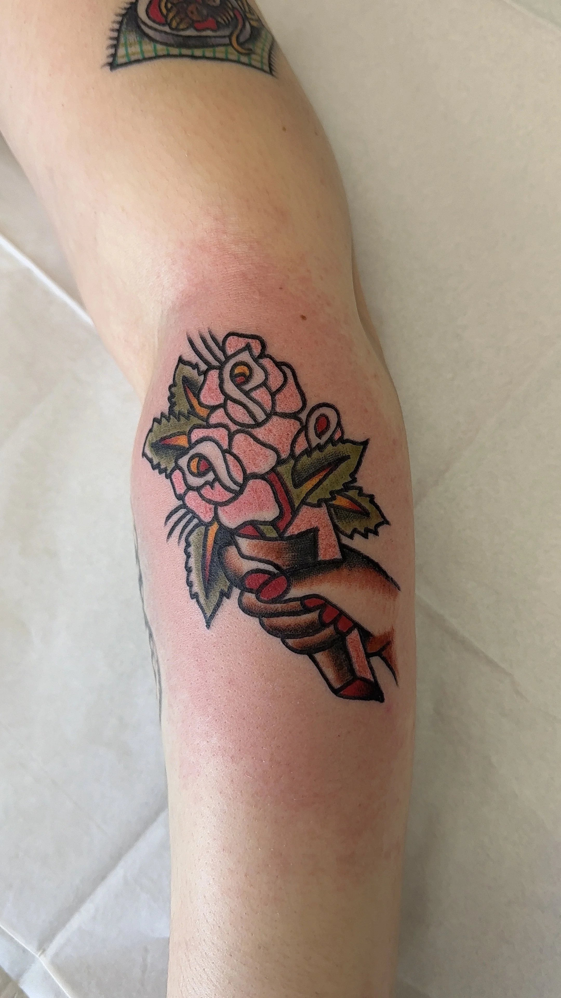 Tattoo of a fist gripping a bouquet of pink roses with green leaves, outlined in black and shaded with red, pink, and green on a person's arm.