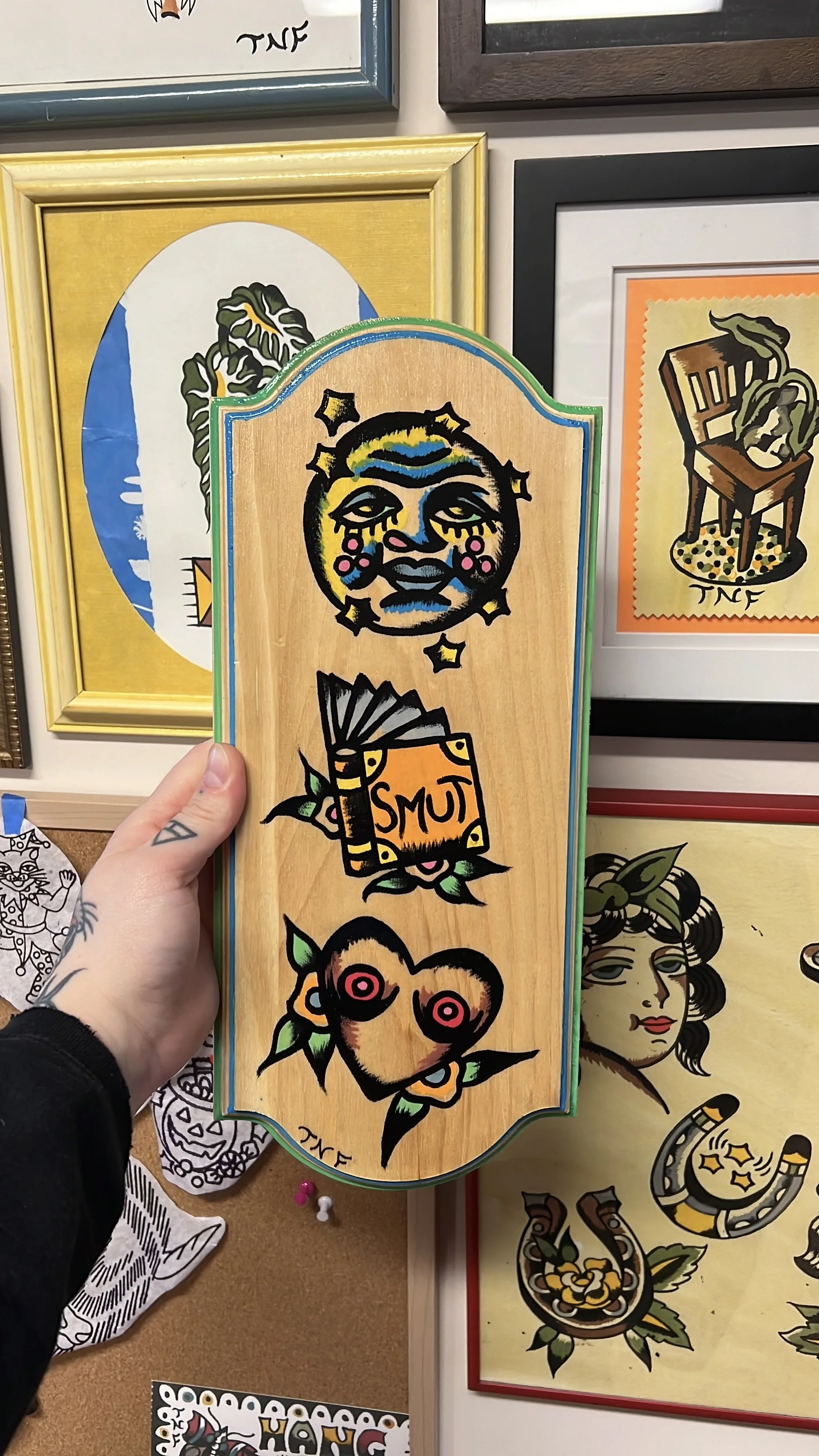 Tattoo-inspired art on a wooden object featuring colorful illustrations of a moon, a book labelled 'smut' and some boobs in the shape of a heart