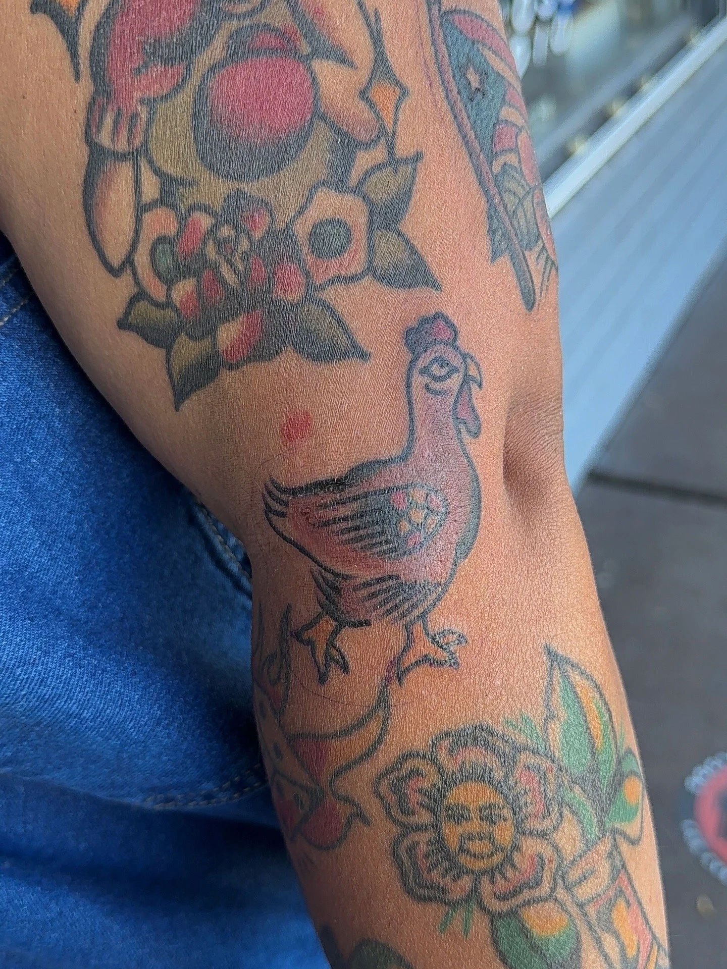 A tattoo on a person's arm featuring a colorful rooster with a flower next to it and other decorative elements.