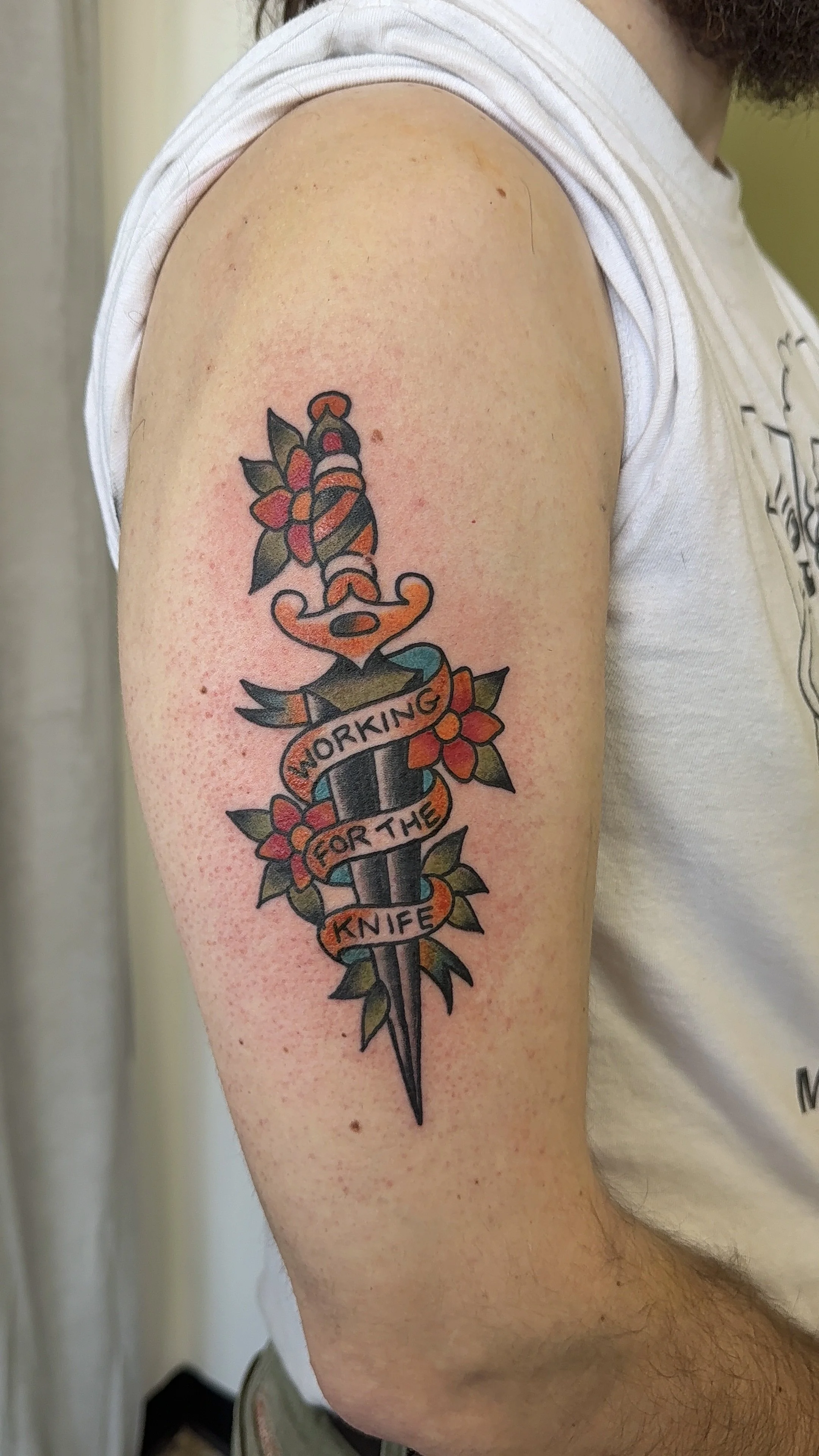 Tattoo of a dagger with flowers wrapped around it and a banner that reads 'WORKING FOR THE KNIFE' on a person's upper arm.