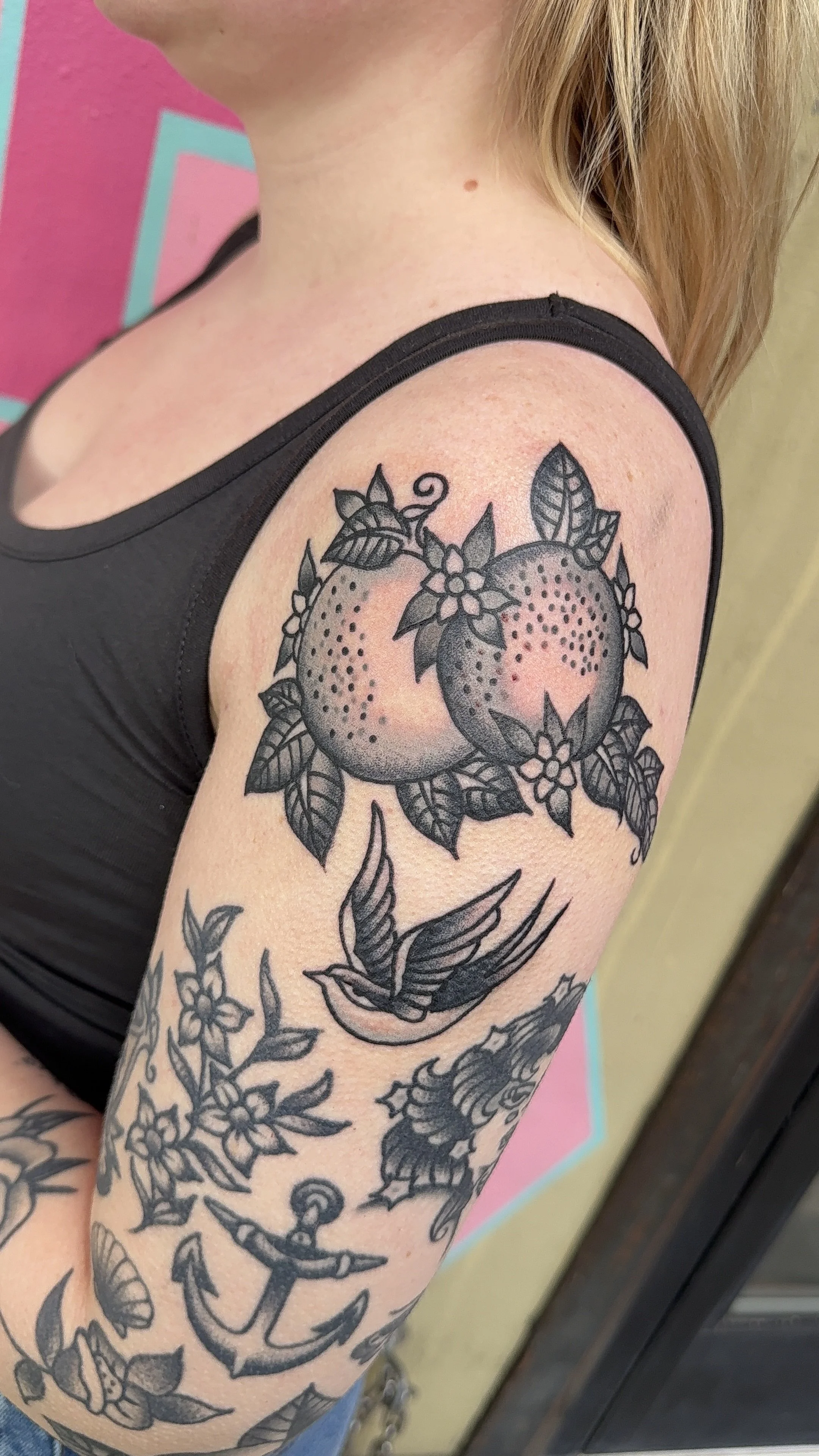 Tattoo of two oranges with leaves and flowers on a person's upper arm and shoulder, black ink outline as well as a swallow bird tattoo in American traditional style