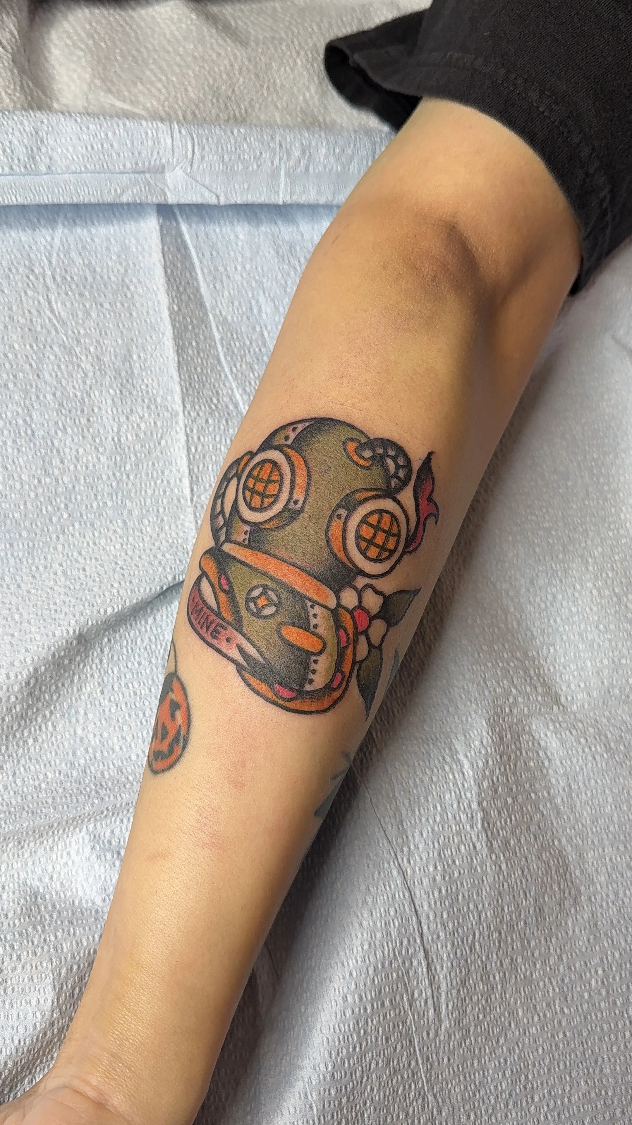 Colorful tattoo of a scuba helmet on a person's forearm, with orange, black, and green details. Tattoo inspired by Hayley Williams album Ego Death at a Bachelorette Party. 