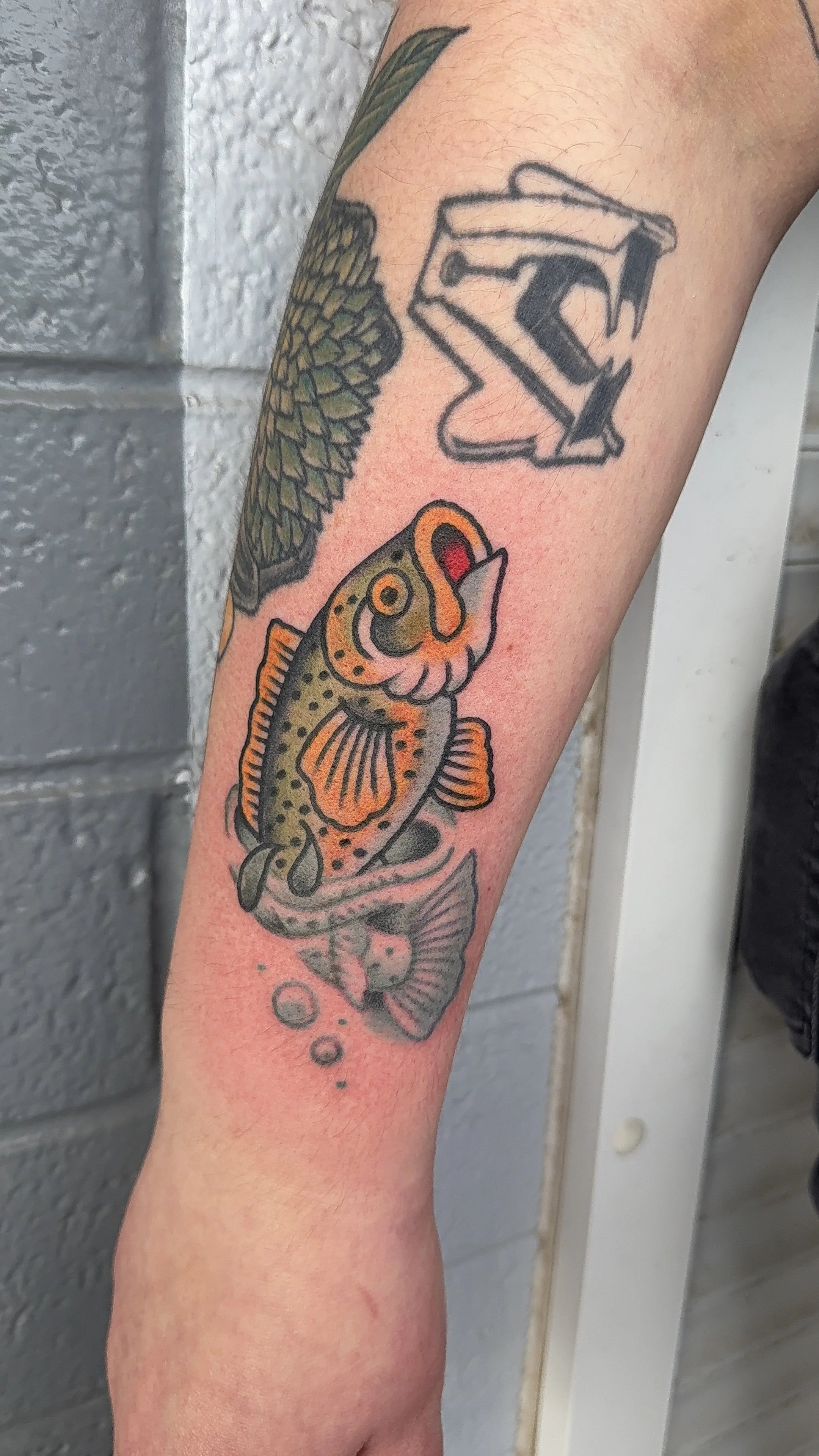 A colorful fish tattoo on a person's forearm, depicting a fish with orange and yellow spots, swimming above bubbles and a small fish, with behind it a black and white tattoo of a house with a chimney.