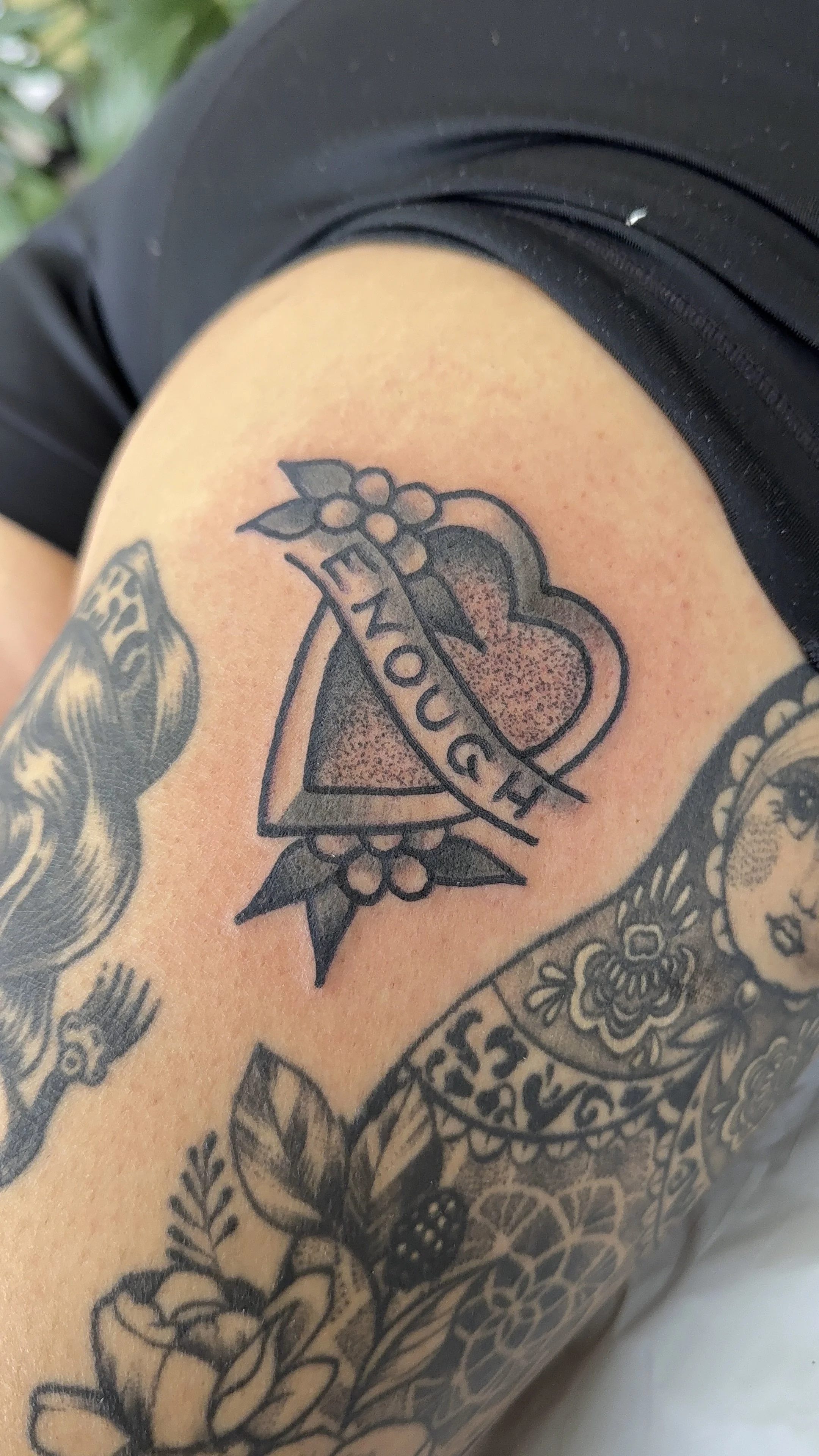 Tattoo of a heart with a banner reading 'ENOUGH' and floral designs on a person's leg.