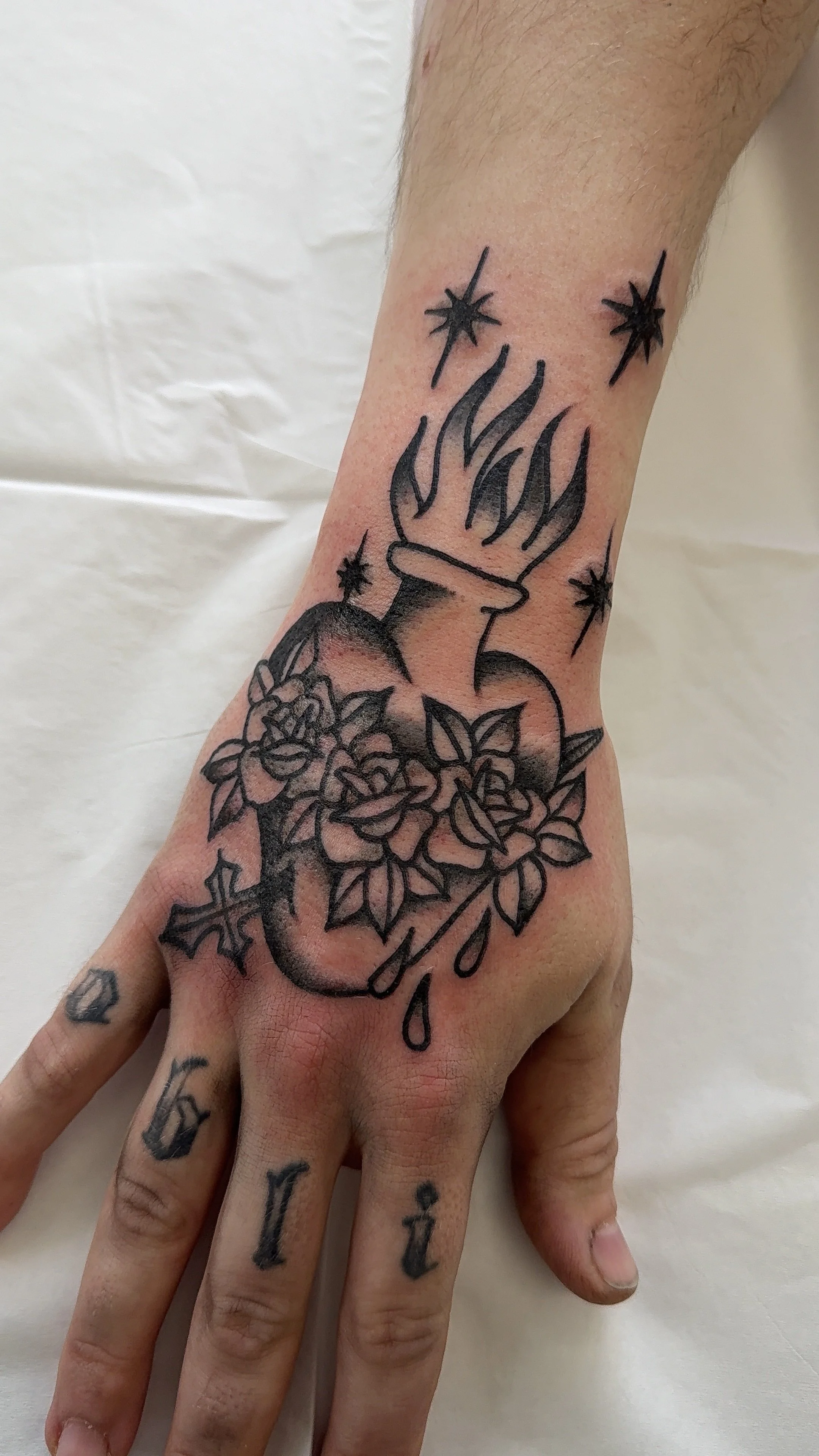 Tattoo of a flaming sacred heart, flowers, and stars on a person's forearm and hand.