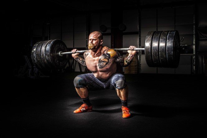 Man with tattoos performing a barbell squat in a gym.