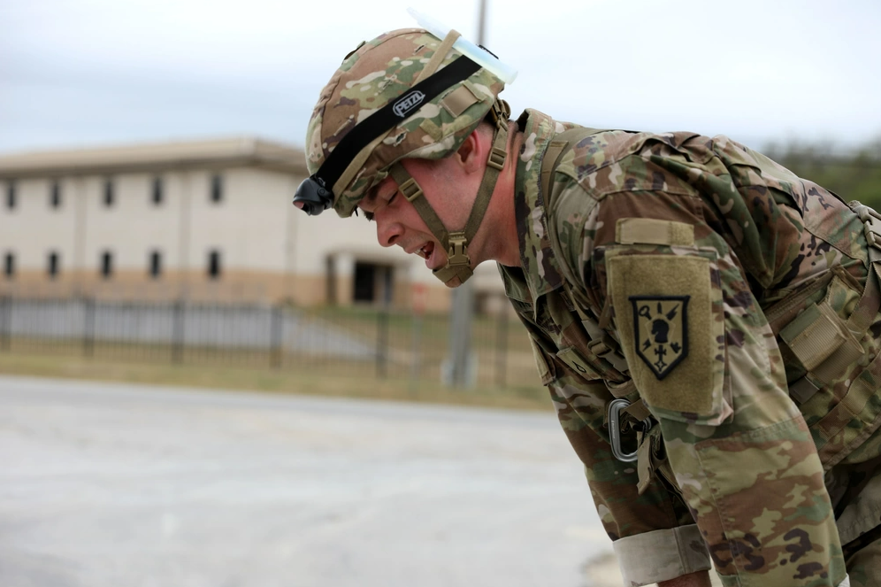 A soldier in camouflage uniform and helmet bending over outdoors, with a building in the background.