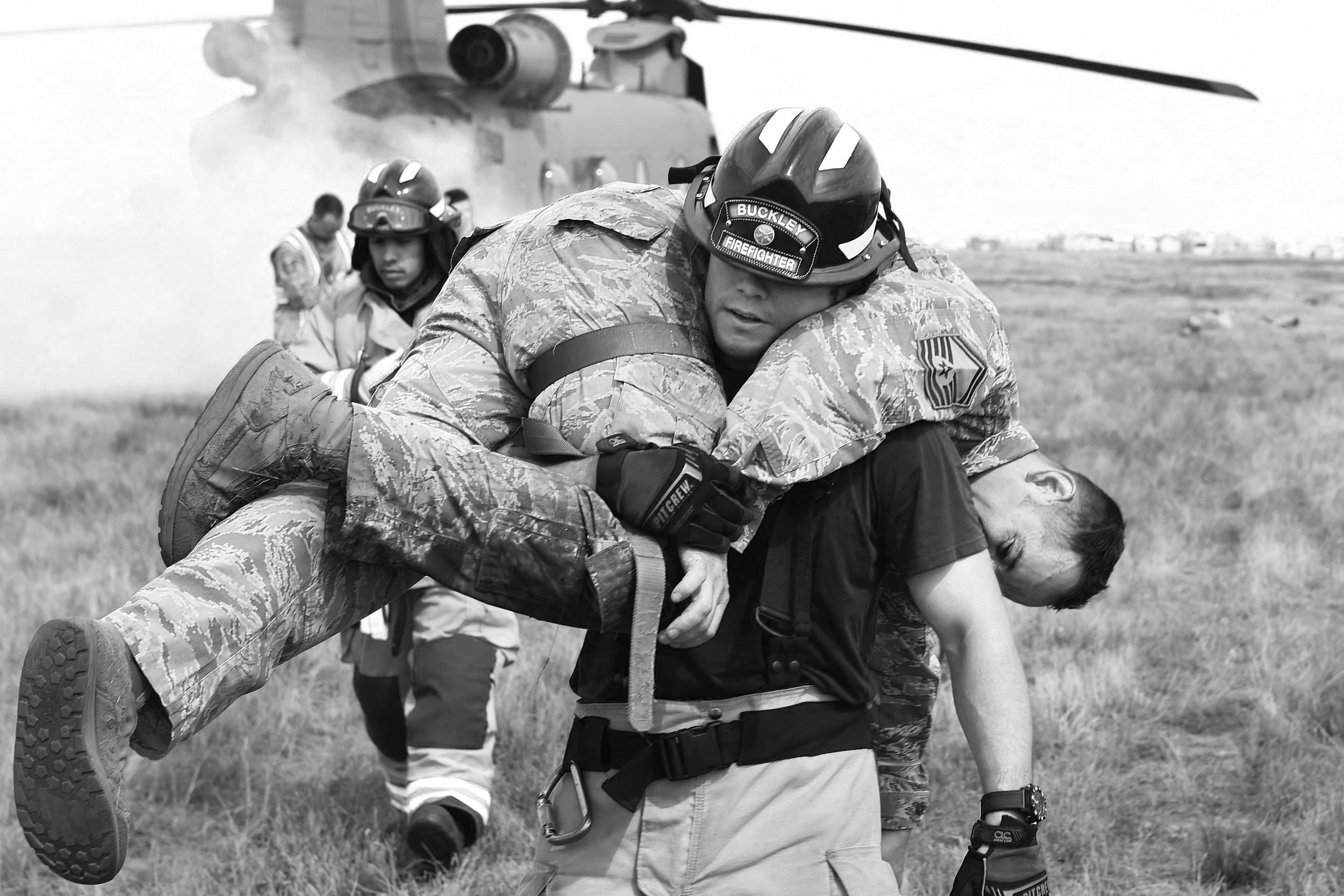 Firefighter carrying an airman during a rescue training exercise, with a helicopter in the background and emergency personnel assisting nearby.