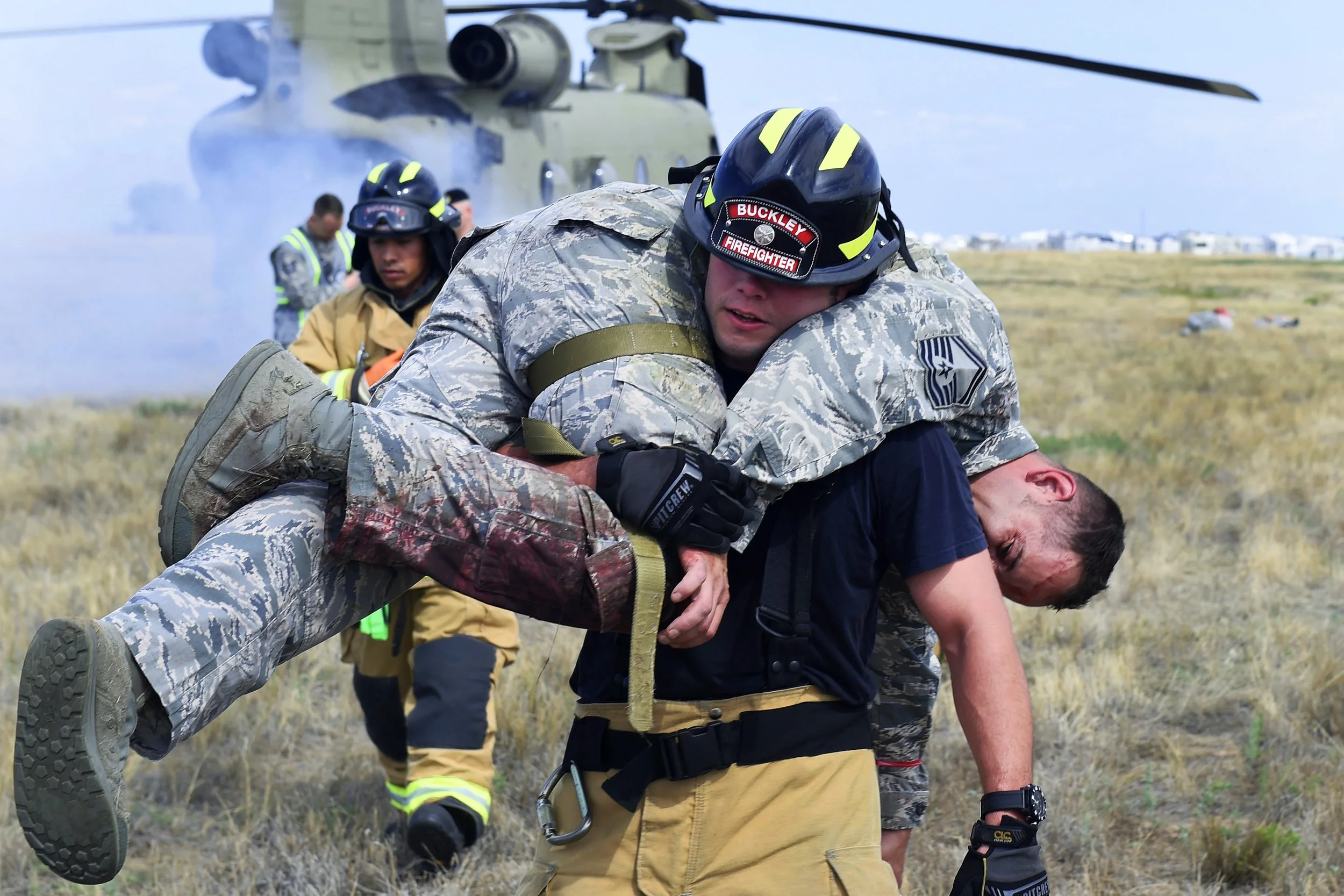 A firefighter carrying a soldier over their shoulder in a field with a helicopter in the background.