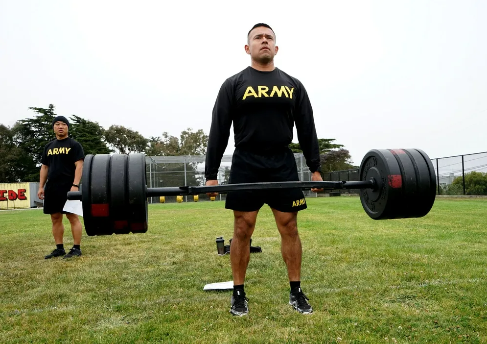 Two army personnel in uniform on a grassy field, one lifting a barbell with weights, the other observing, with trees and a fence in the background.
